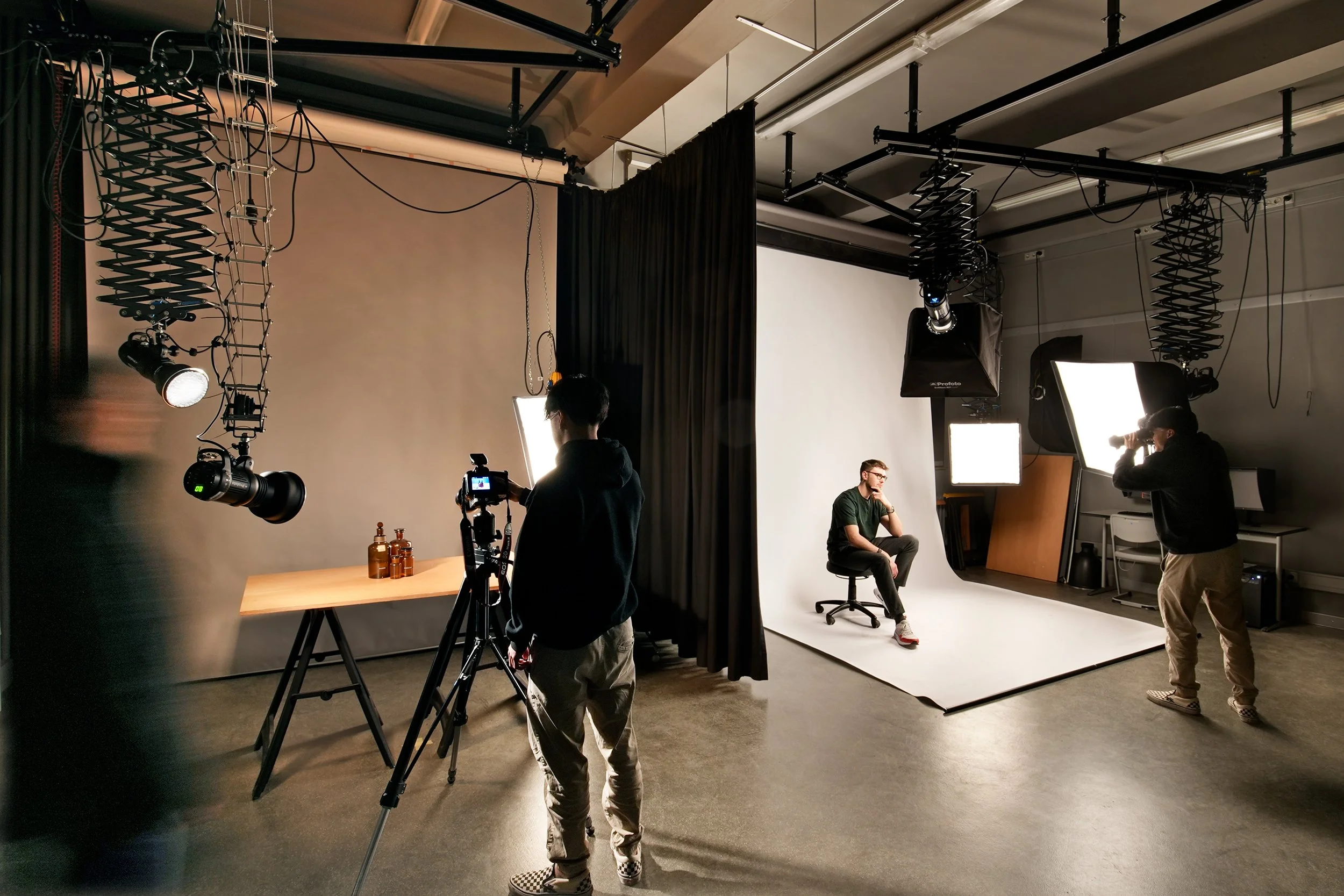 Eine Fotografieszene eines Fotostudios mit einem Model, das auf einem weißen Hintergrund sitzt, und mehreren Personen bei der Arbeit mit Beleuchtung und Kamera. Es gibt Studioausrüstung und und schwarz abgehängte Vorhänge.