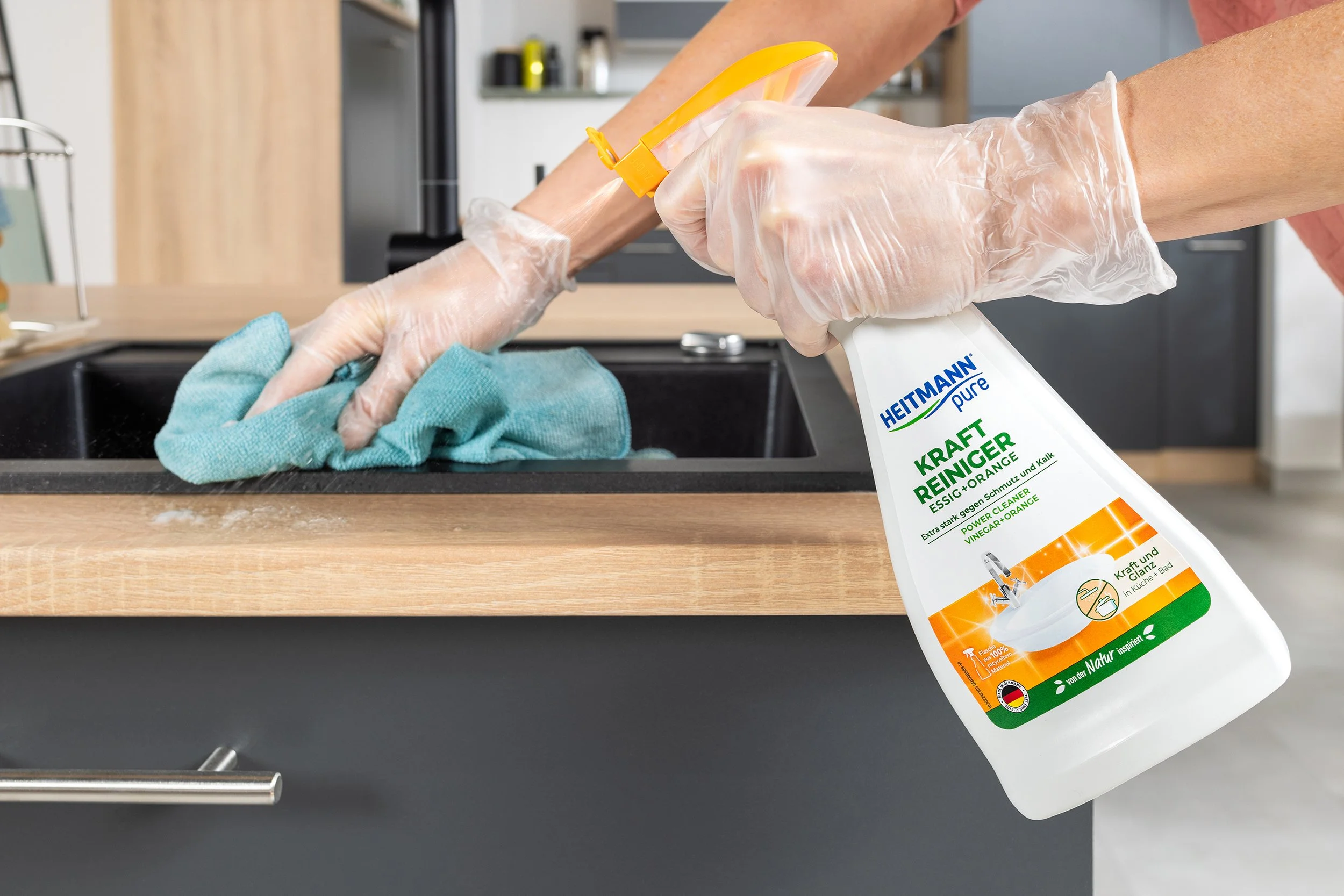 Hand mit einem Gummihandschuh beim Reinigen eines schwarzen Spülbeckens, Wassersprüher mit der Aufschrift 'Heitmann Pure Kraft Reiniger' in die Kamera gehalten.
