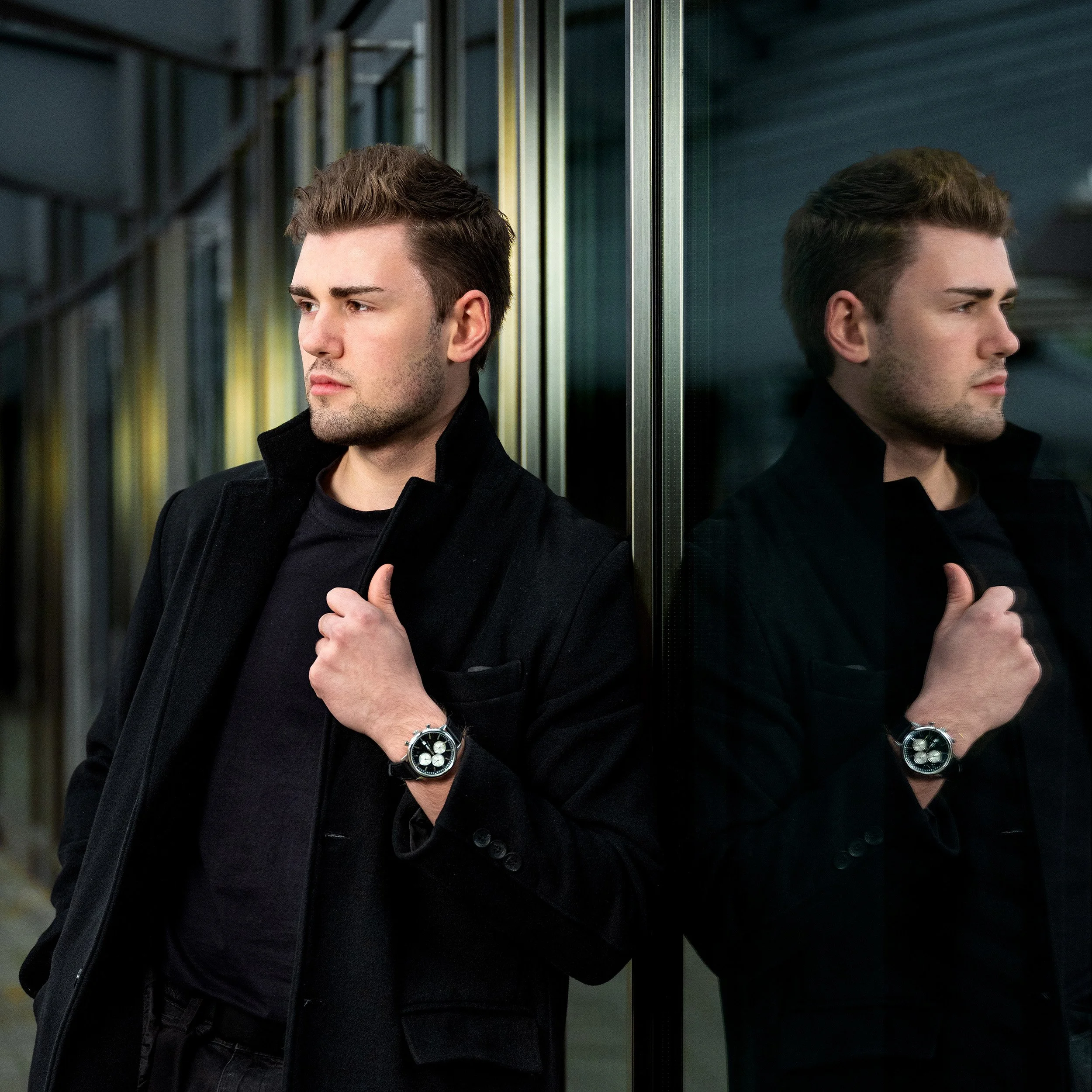 Ein junger Mann mit dunklem Haar trägt eine schwarze Jacke und eine Armbanduhr und steht neben einem Spiegel, der seine Reflexion zeigt.