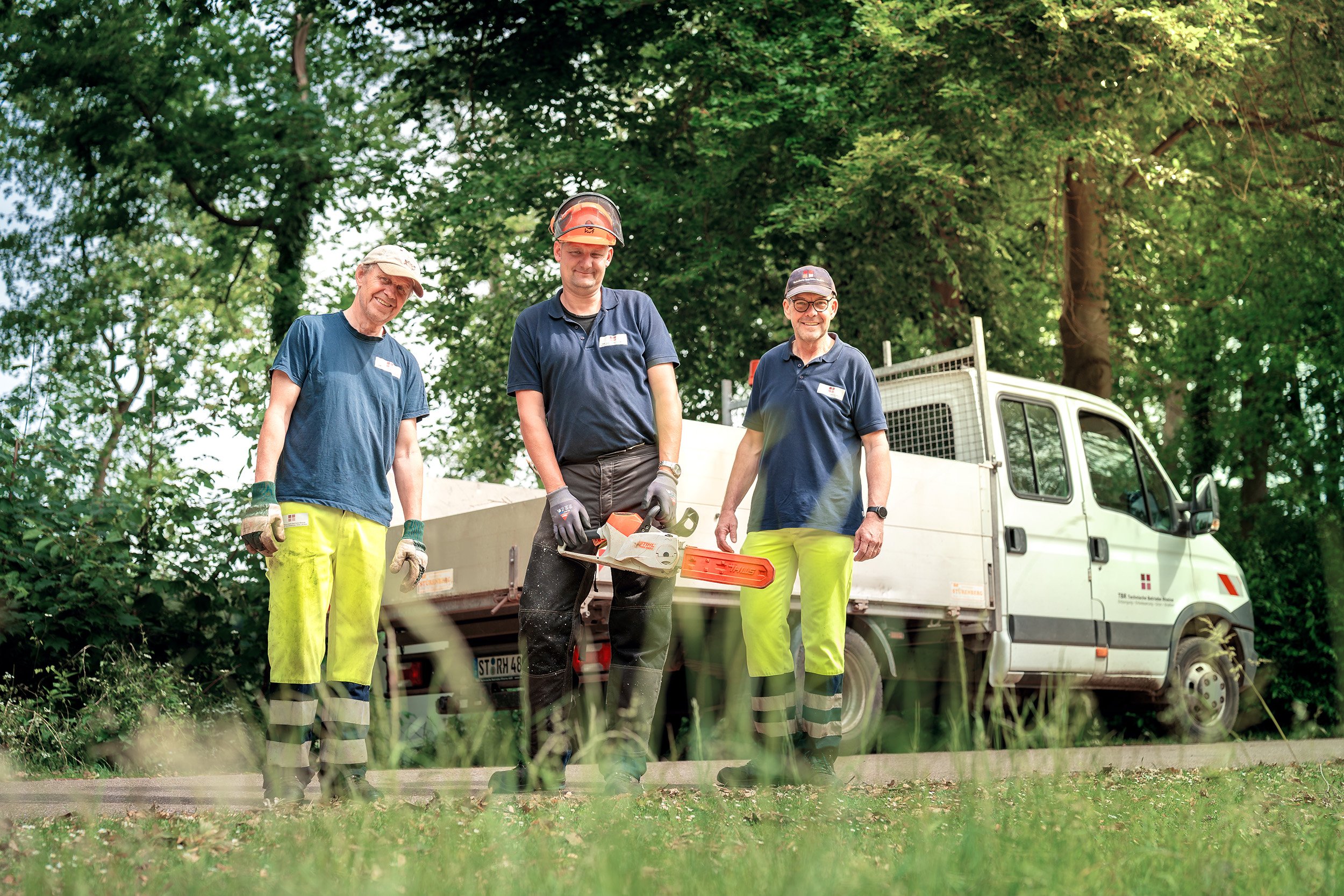 Drei Männer in Arbeitskleidung stehen vor einem weißen Arbeitstransporter, umgeben von Bäumen, im Freien.