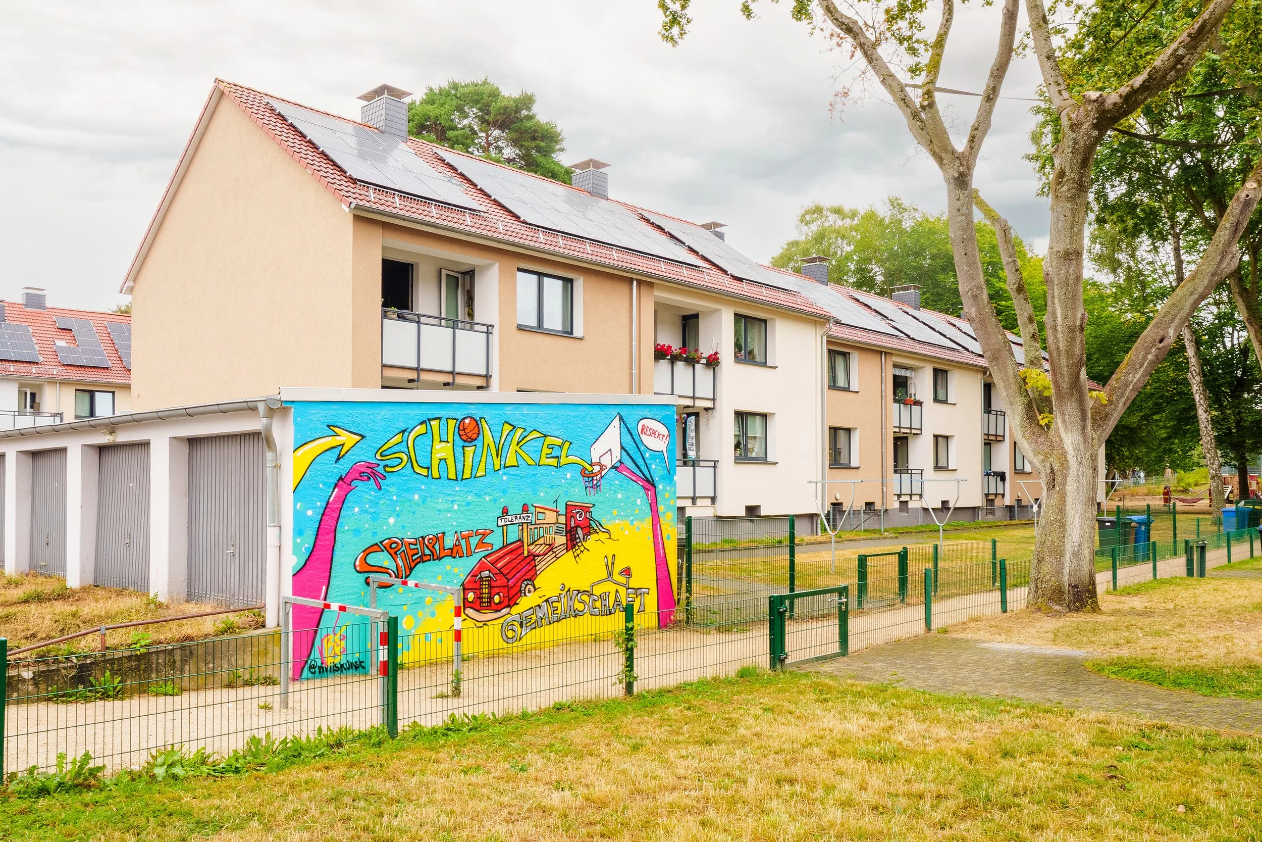 Mehrstöckiges Mehrfamilienhaus mit roten Dachziegeln und Solarpanelen auf dem Dach, umgeben von Bäumen und einem Garten mit Spielplatz, umfriedet von einem grünen Zaun.