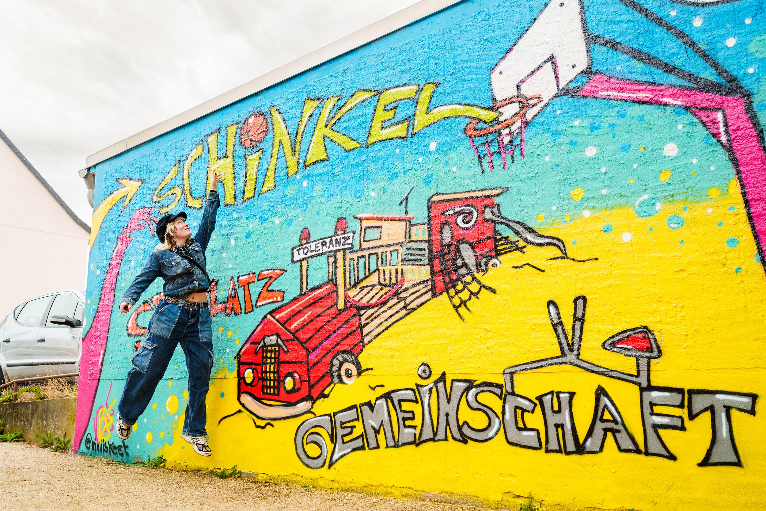 Eine Person steht vor einer bunten Wand mit Graffiti-Kunst und streckt die Hand nach dem Namen 'SchanKEL' aus.