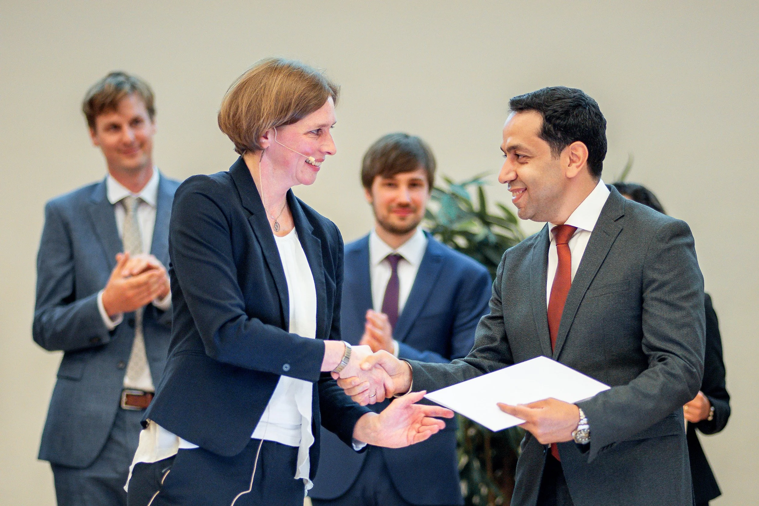 Zwei Menschen schütteln sich die Hände bei einer formellen Veranstaltung, im Hintergrund applaudierende weitere Personen.