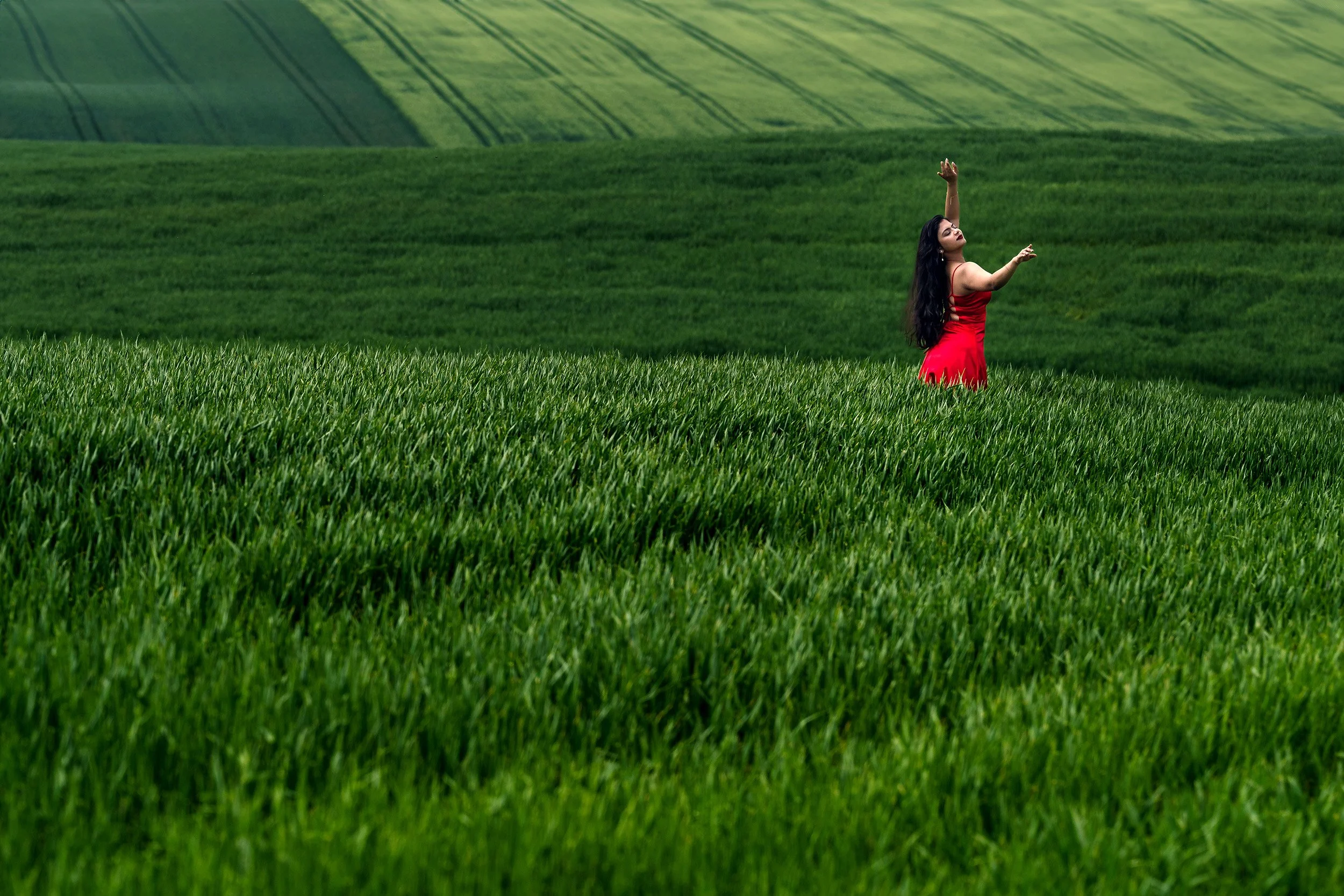 Eine Frau in einem roten Kleid steht in einem grünen Feld mit Hügeln im Hintergrund, die mit Streifen von Pflanzen bedeckt sind.