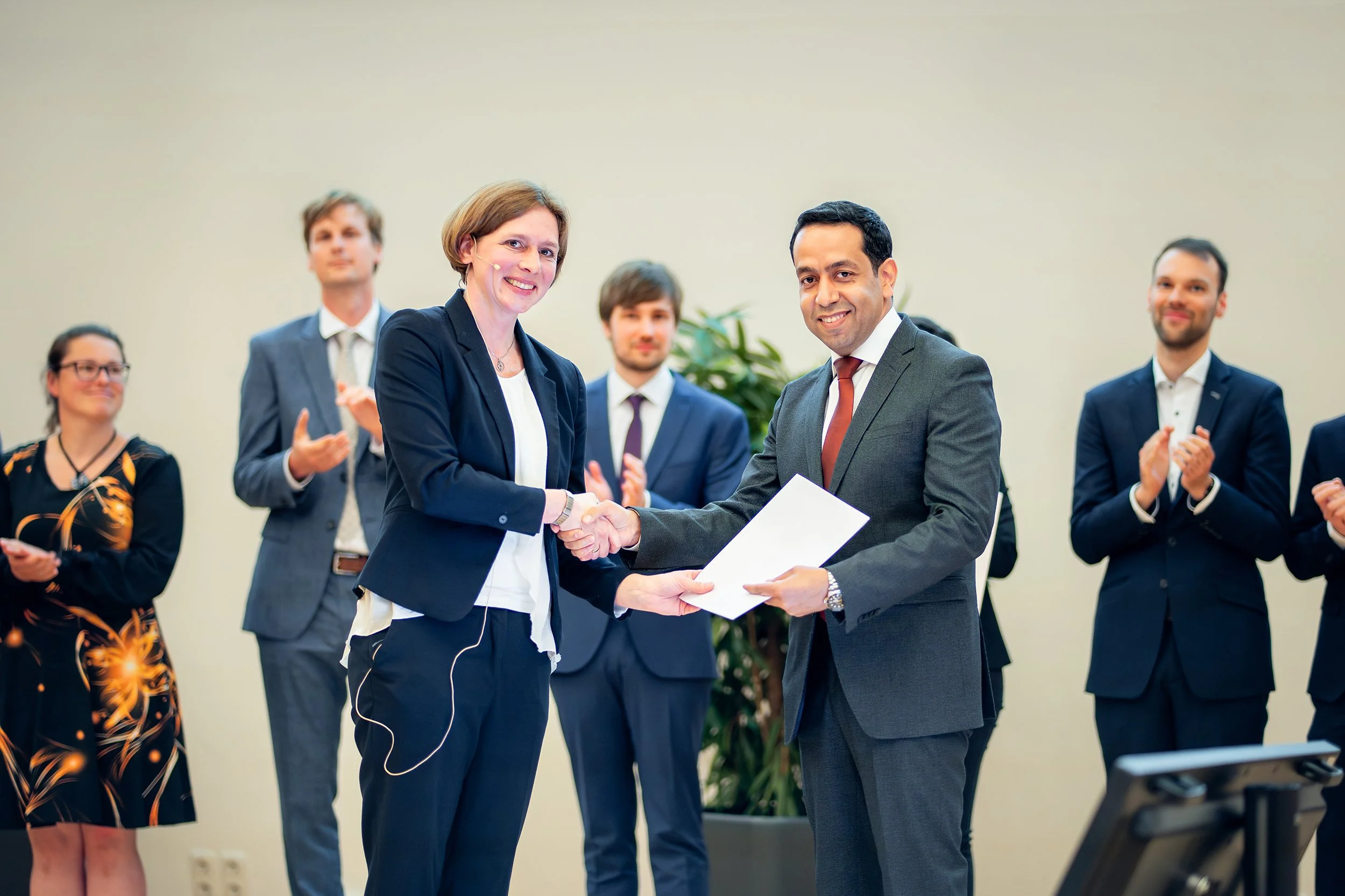 Zwei Menschen schütteln sich bei einer formellen Zeremonie oder einem Geschäftstreffen die Hand, um eine Vereinbarung oder einen Vertrag zu besiegeln. Im Hintergrund applaudieren und beobachten weitere Menschen die Szene.