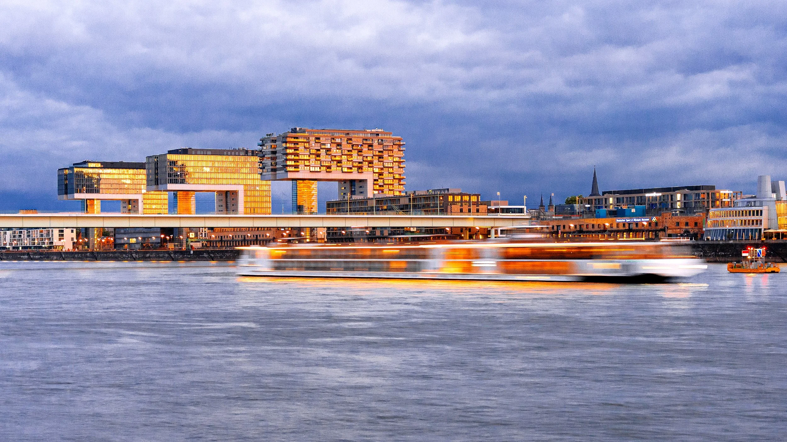 Moderne Gebäude am Flussufer bei Sonnenuntergang, reflektierende Fenster, ein fahrendes Boot im Wasser.