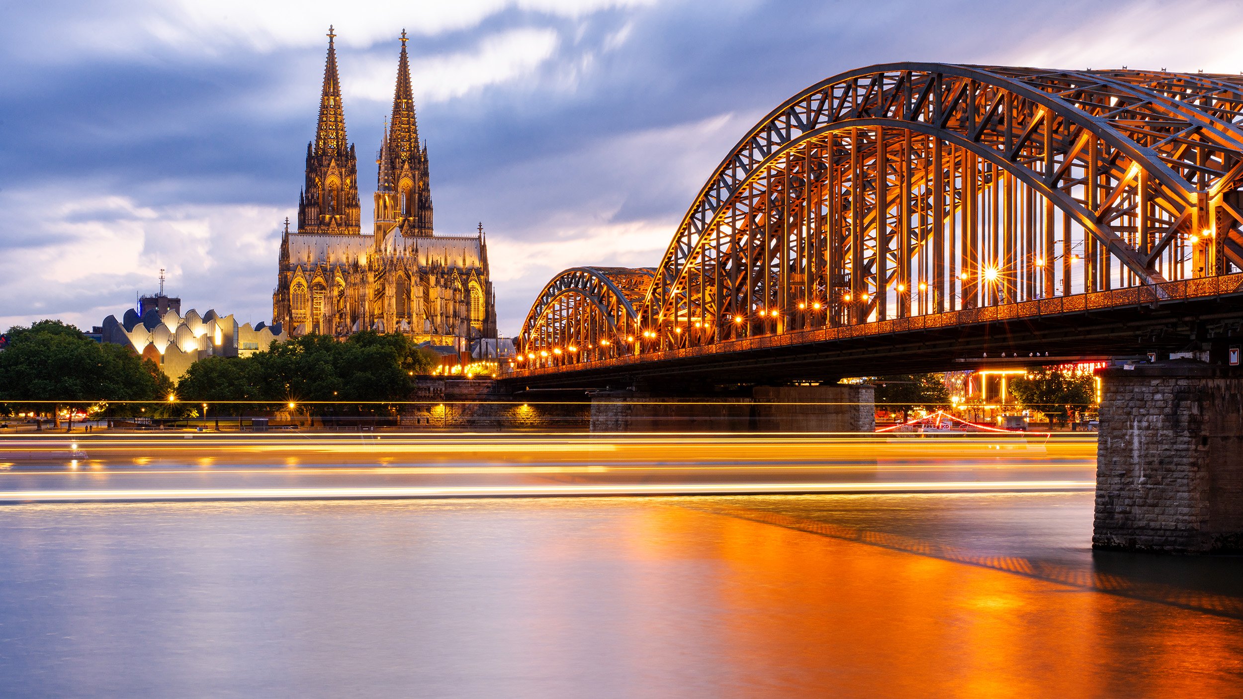 Blick auf die Kölner Dom und die Hohenzollernbrücke bei Sonnenuntergang, mit beleuchteter Brücke und Wasserreflexionen.