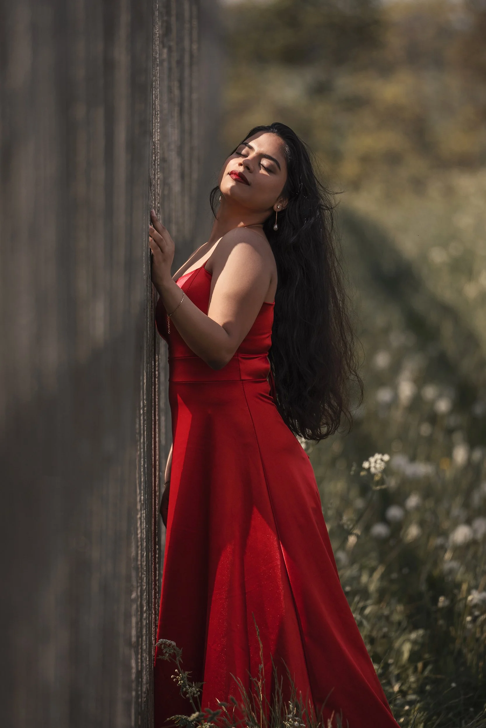 Eine Frau in einem roten Kleid steht an einer Holzwand im Freien, mit geschlossenen Augen und einem entspannten Ausdruck, während sie Sonnenlicht auf ihr Gesicht fällt.