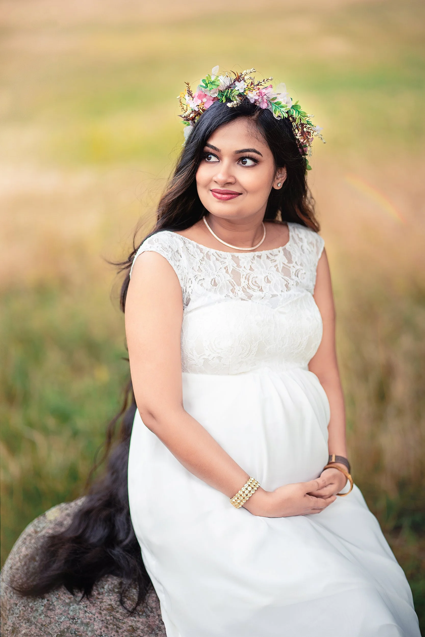 Junge Frau im weißen Kleid mit Blumenkrone sitzt im Grünen, hält ihre Hände vor ihren Bauch, hat lange dunkle Haare, trägt Schmuck und schaut nach rechts.