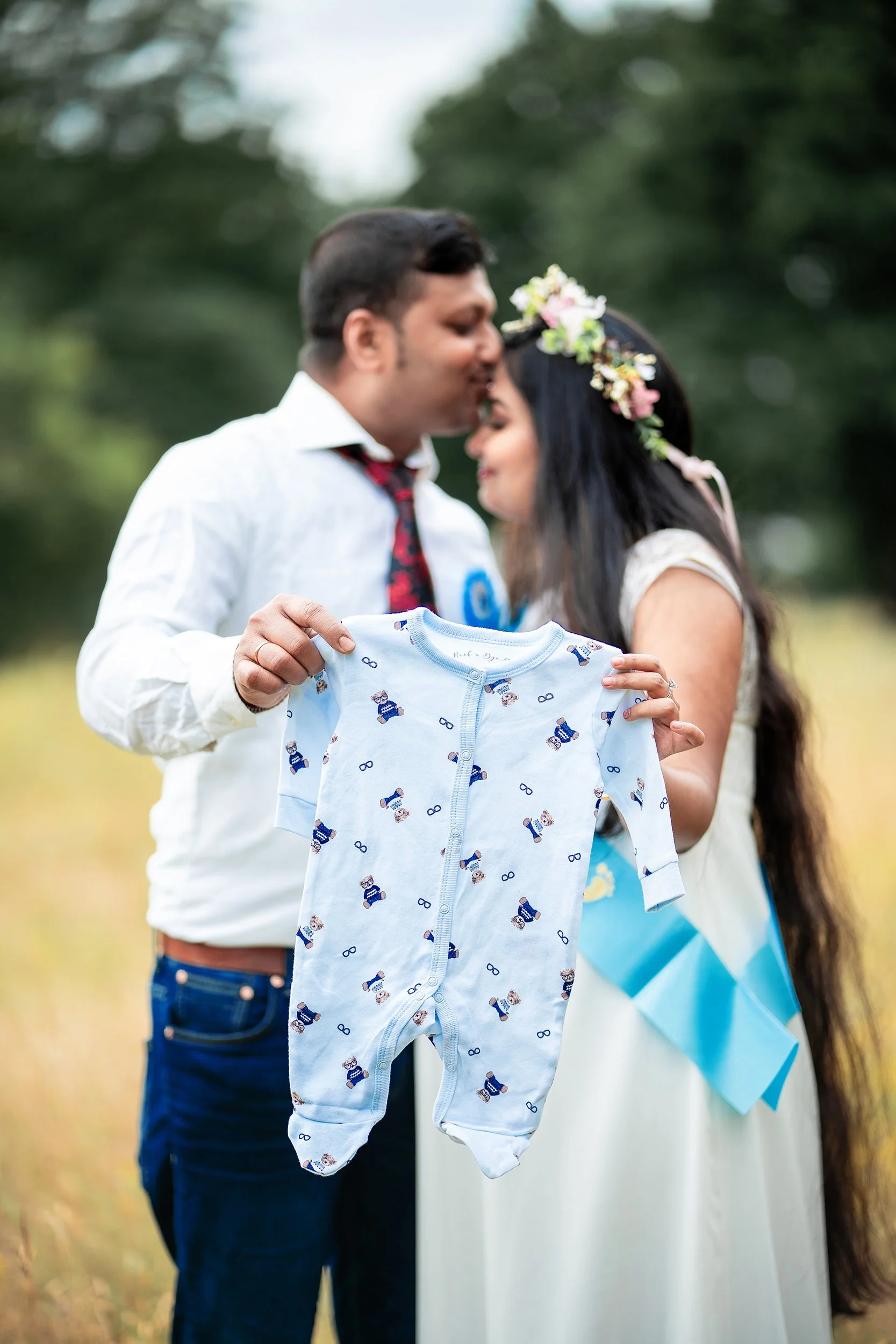 Ein Paar, wahrscheinlich ein Brautpaar, hält eine Baby-Strampler in der Hand, während sie im Freien in einer grünen, natürlichen Umgebung stehen.