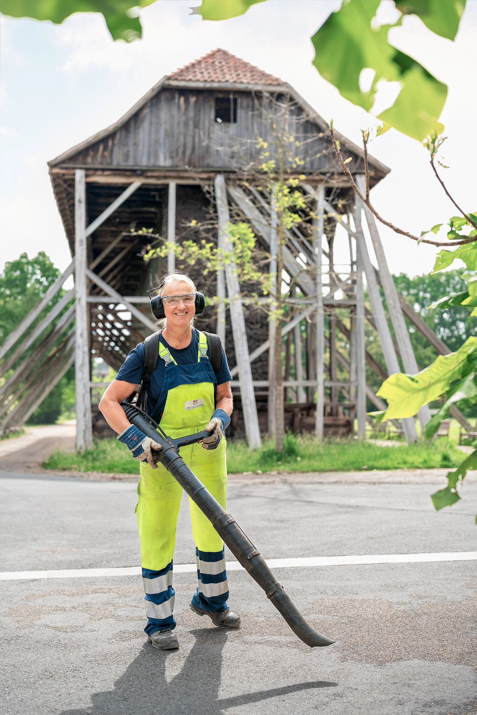 Eine Frau in Arbeitshose und Schutzbrille benutzt einen Laubbläser vor einem alten Holzhaus.