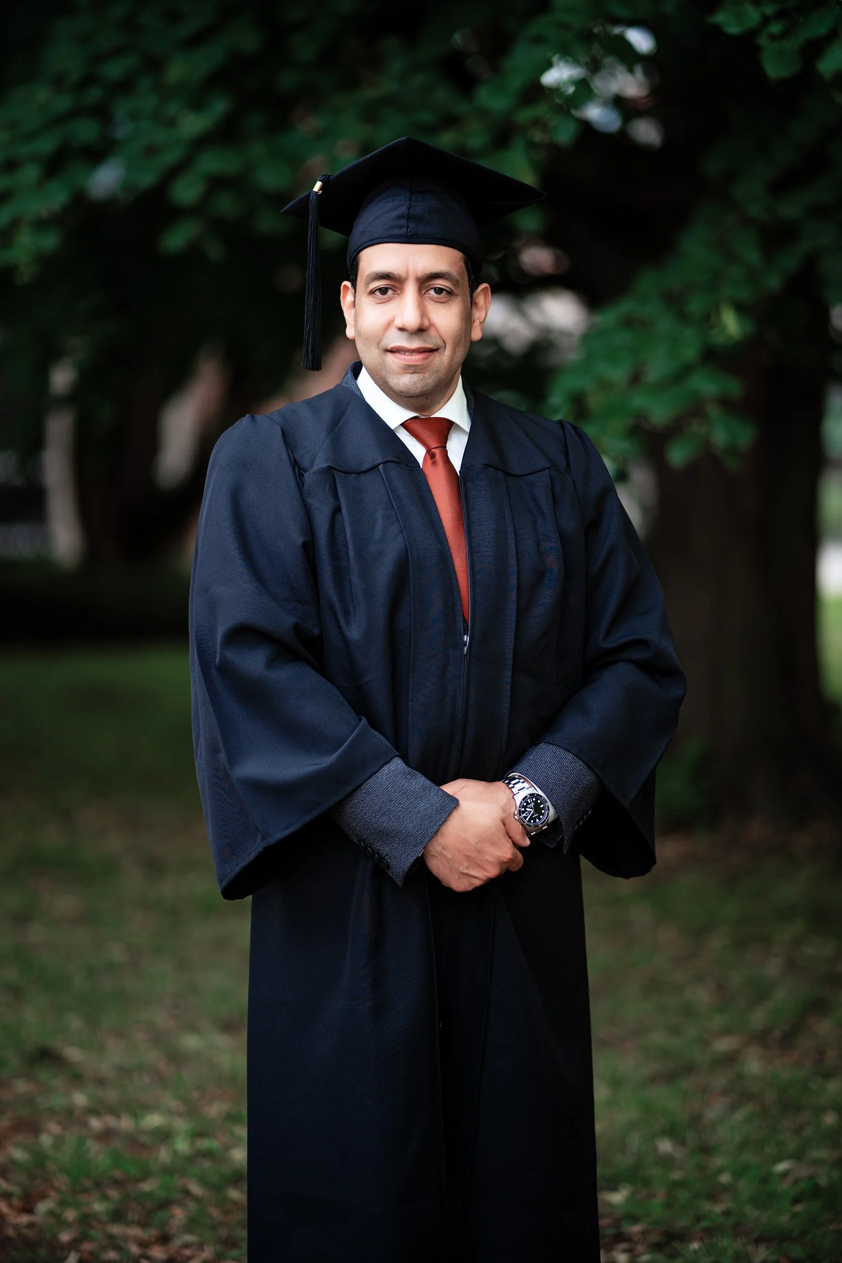 Ein Mann in Graduierungskleidung mit Doktorhut steht im Freien vor grünen Bäumen.