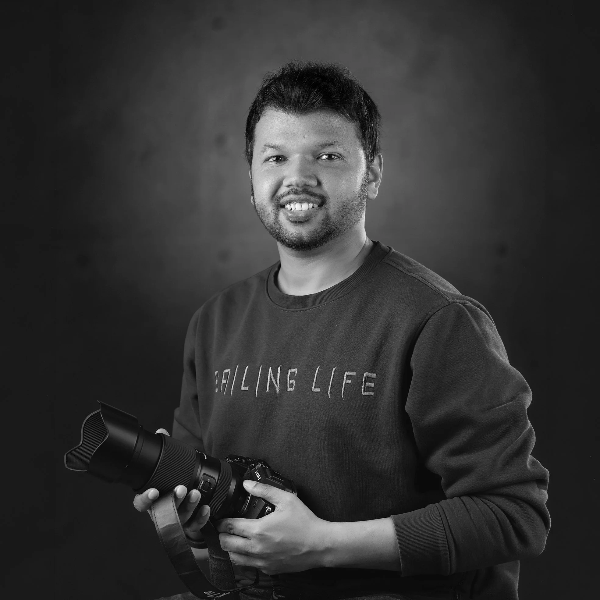Schwarz-weiß-Fotografie eines jungen Mannes mit dunklem Haar und Bart, der einen Videokamera festhält. Er trägt ein dunkles Oberteil mit geschriebenem Text. Der Hintergrund ist unscharf dunkel.