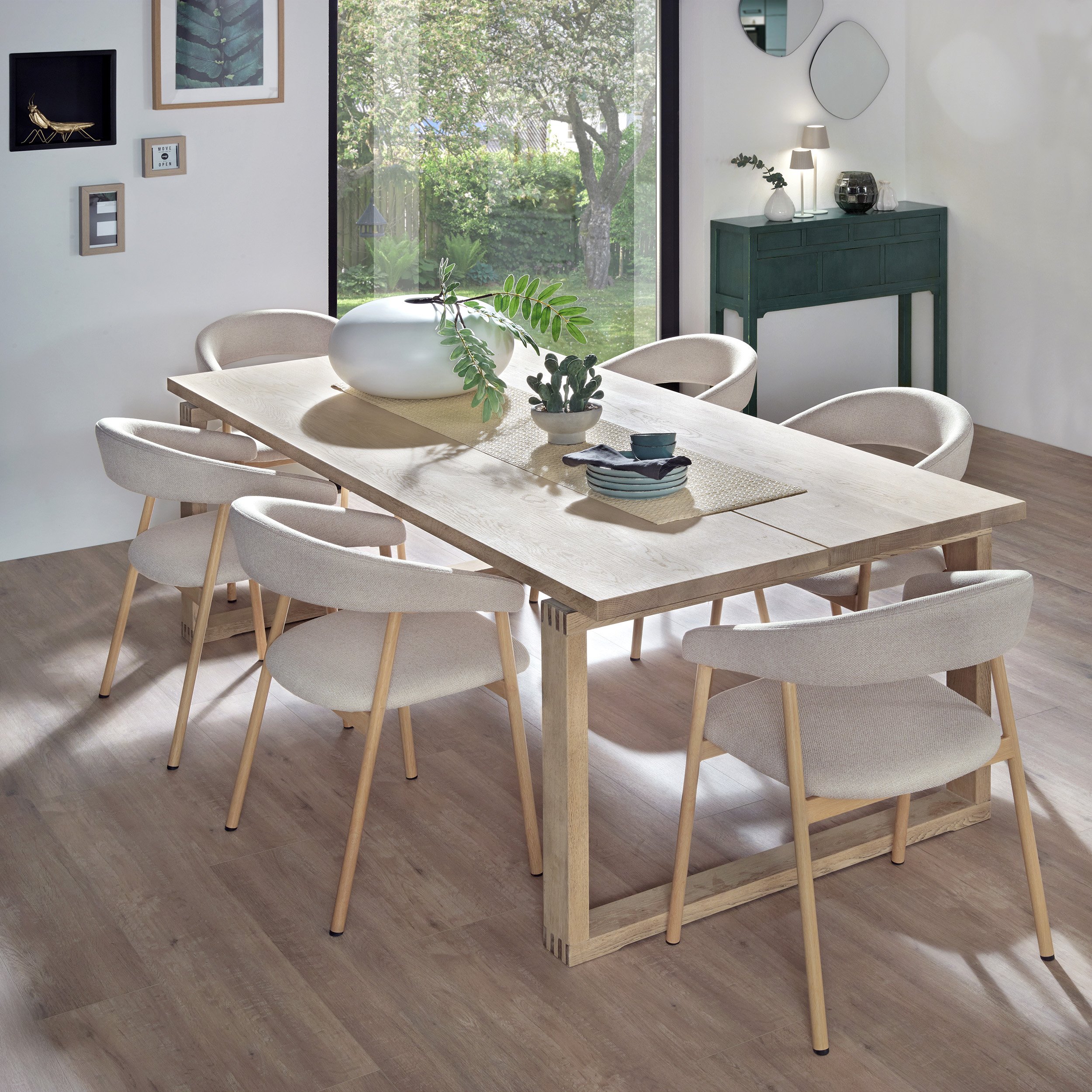 Ein heller Esstisch aus Holz mit weißen Polsterstühlen im modernen Esszimmer. Auf dem Tisch befindet sich eine weiße Vase mit grünen Zweigen. Im Hintergrund ist eine große Fensterfront mit Blick auf einen Garten mit Bäumen und grünem Gras. An der Wan