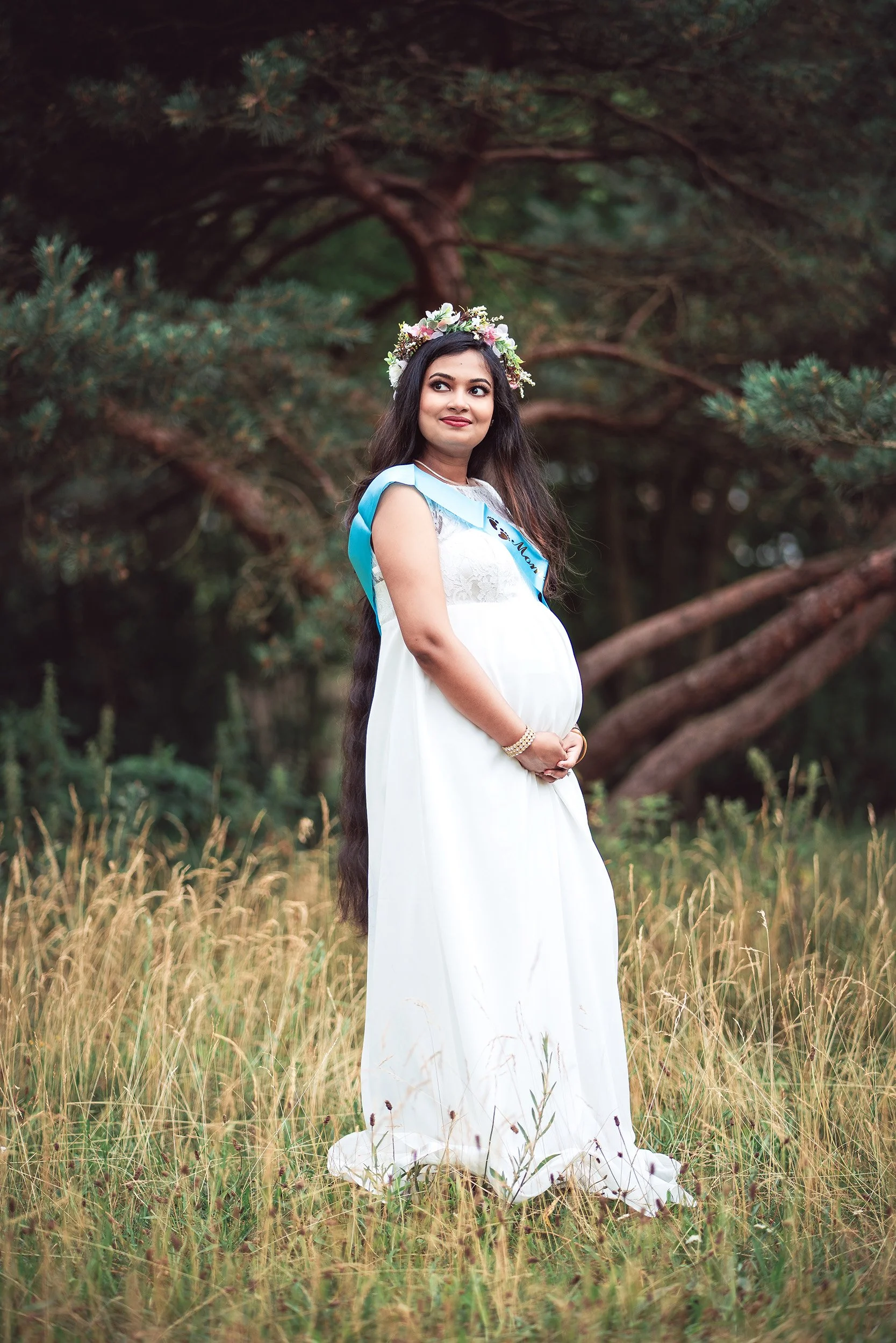 Eine junge Frau in einem weißen Kleid mit einem Blumenkranz auf dem Kopf steht in einem Naturgebiet mit hohen Gräsern und Bäumen im Hintergrund.