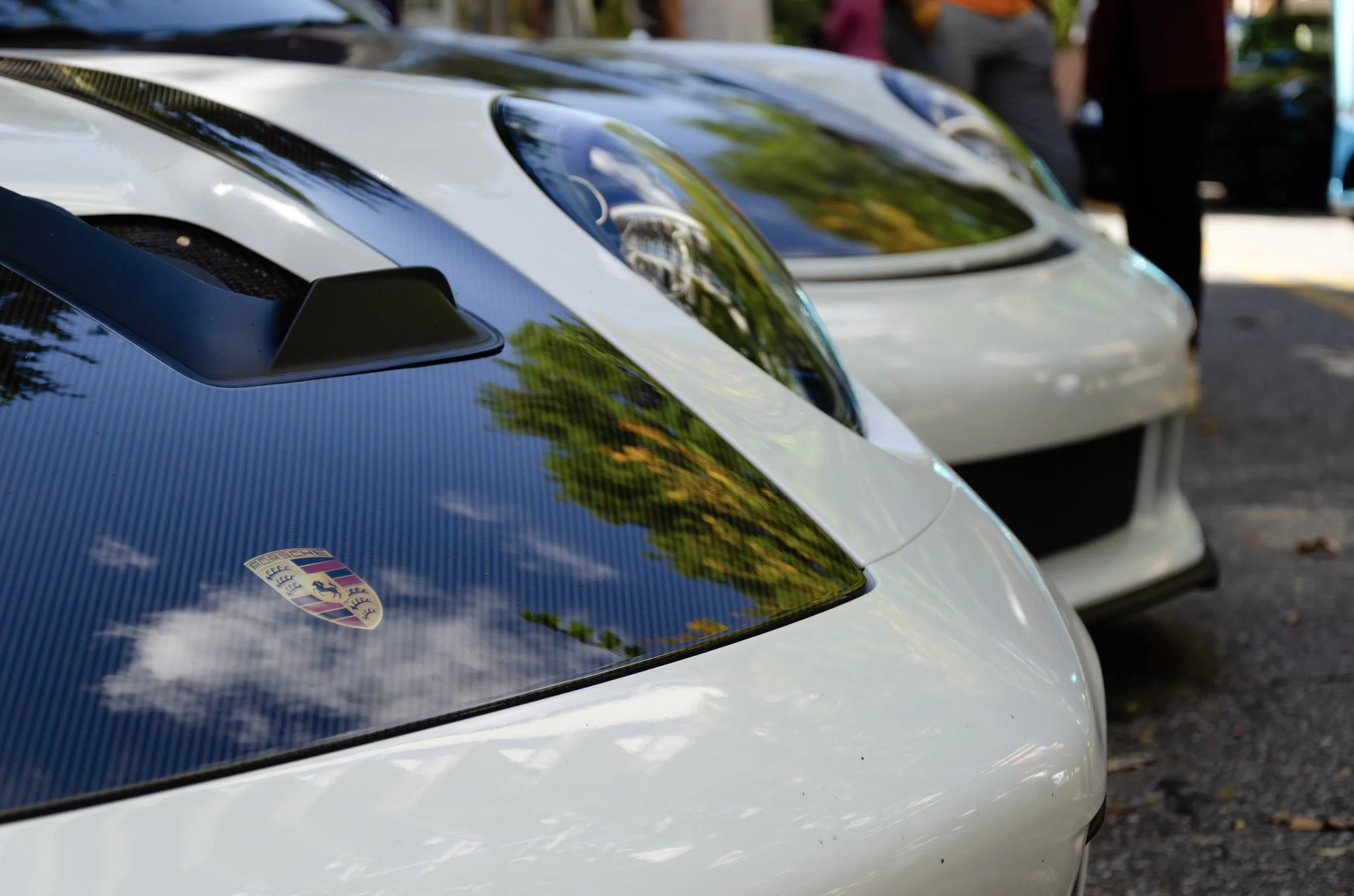 Close-up of the front parts of two  white Porsche sports cars.  The cars feature sleek aerodynamic design and are parked in a row, reflections of trees visible on the car's surfaces.  The cars were a part of an Formula 1 watch party in Buckhead, GA.