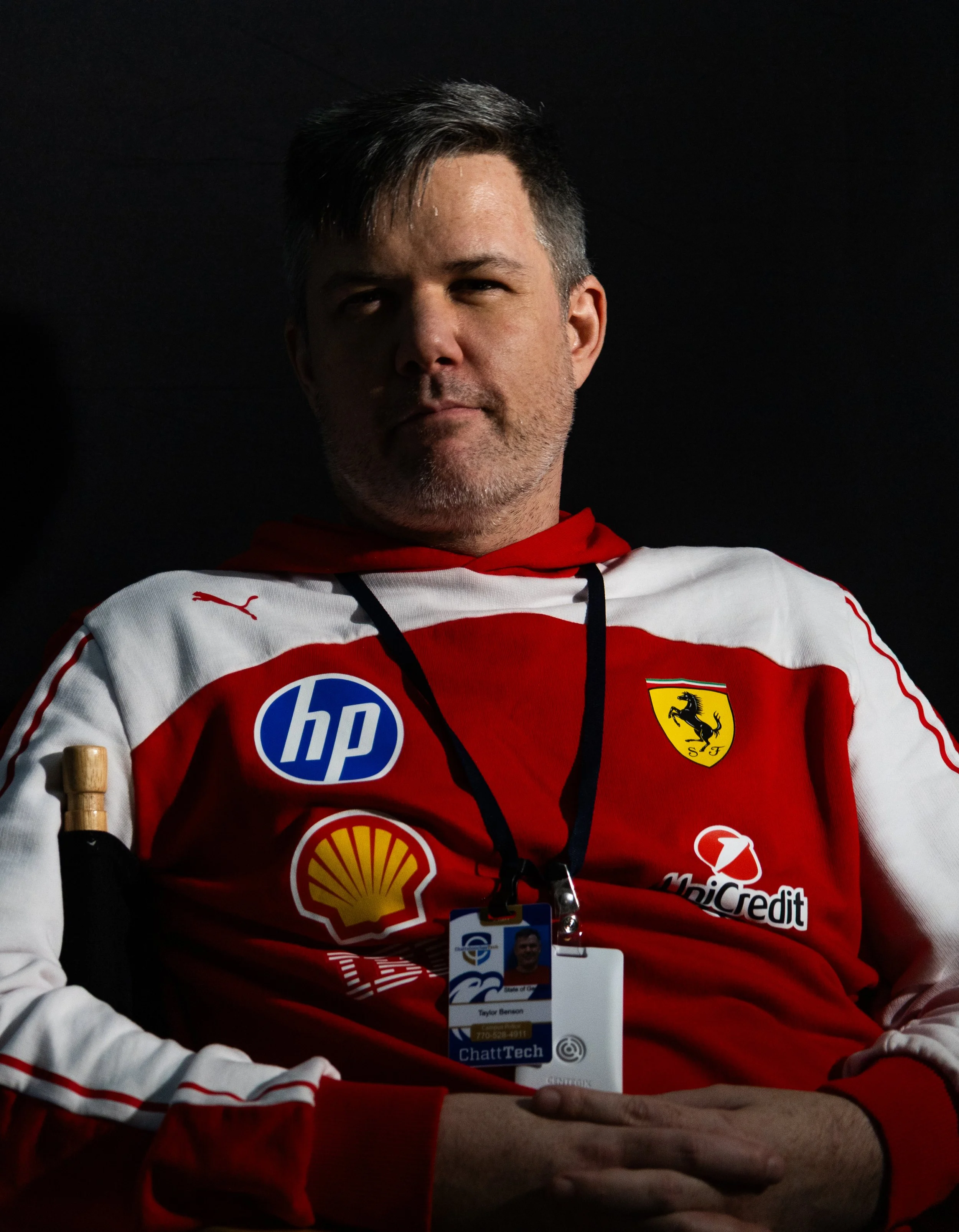 The photographer named Taylor Benson, sitting for a portrait in a photography studio.  The man is wearing a hoodie from the Formula 1 team Scuderia Ferrari, which displays sponsor logos on the clothing that sit on the iconic team color of scarlet red