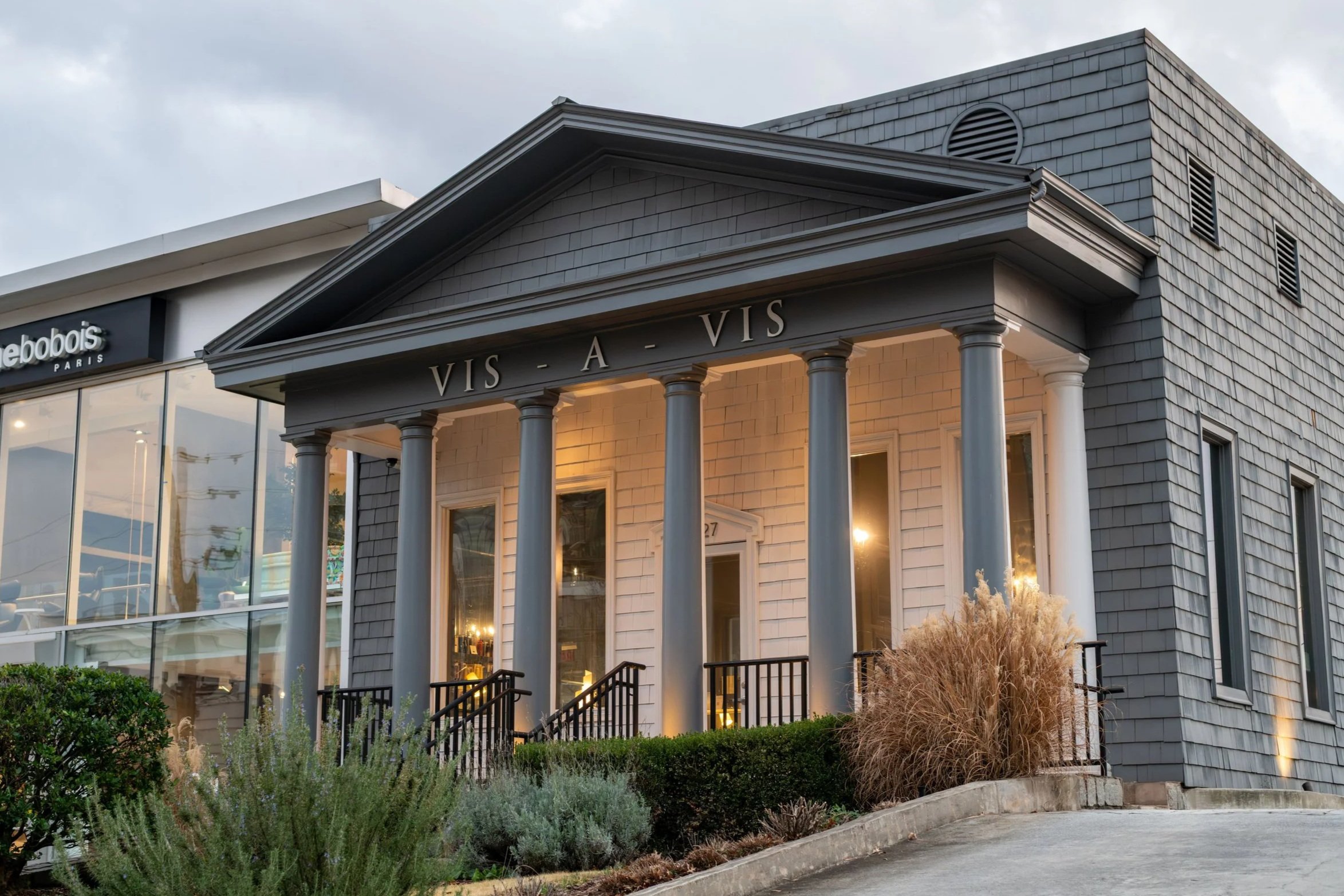 Exterior of a building with classical architecture elements, including tall white columns supporting a triangular pediment with the words 'VIS-A-VIS'. The building has gray shingled siding and large glass windows. Landscaping with bushes and ornamental grasses is visible in front.
