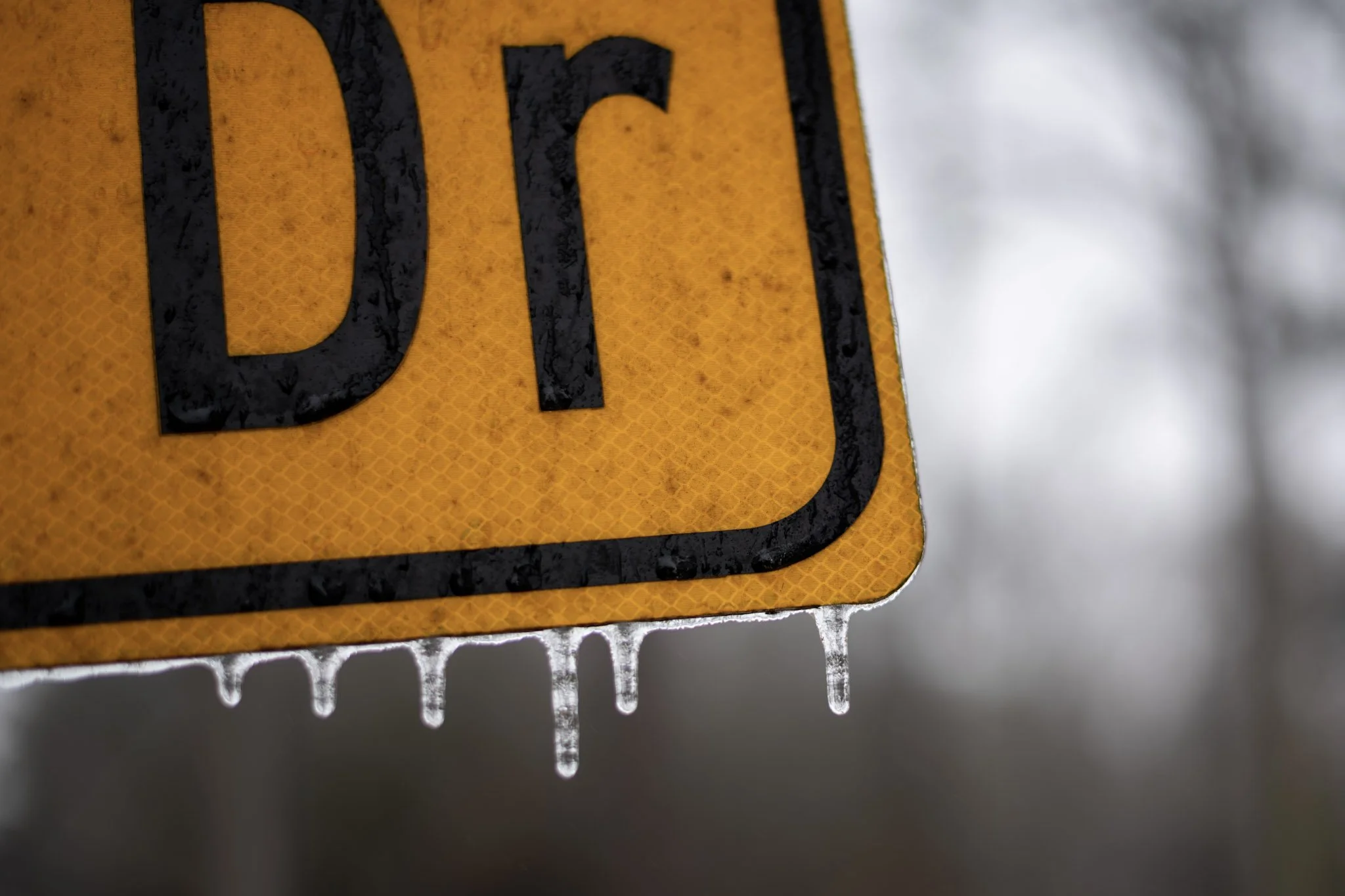 Close-up photo of a yellow caution road sign with black text, frosted with ice and icicles hanging at the bottom, indicating cold weather.  The photo occurred during the Georgia ice storm from January 2026.