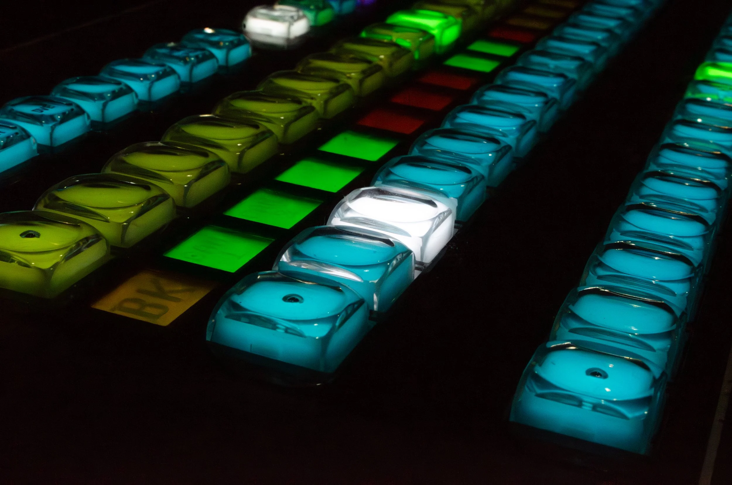 Close-up of illuminated buttons for a Ross video control board arranged in rows, featuring blue, yellow, white, and green colors.