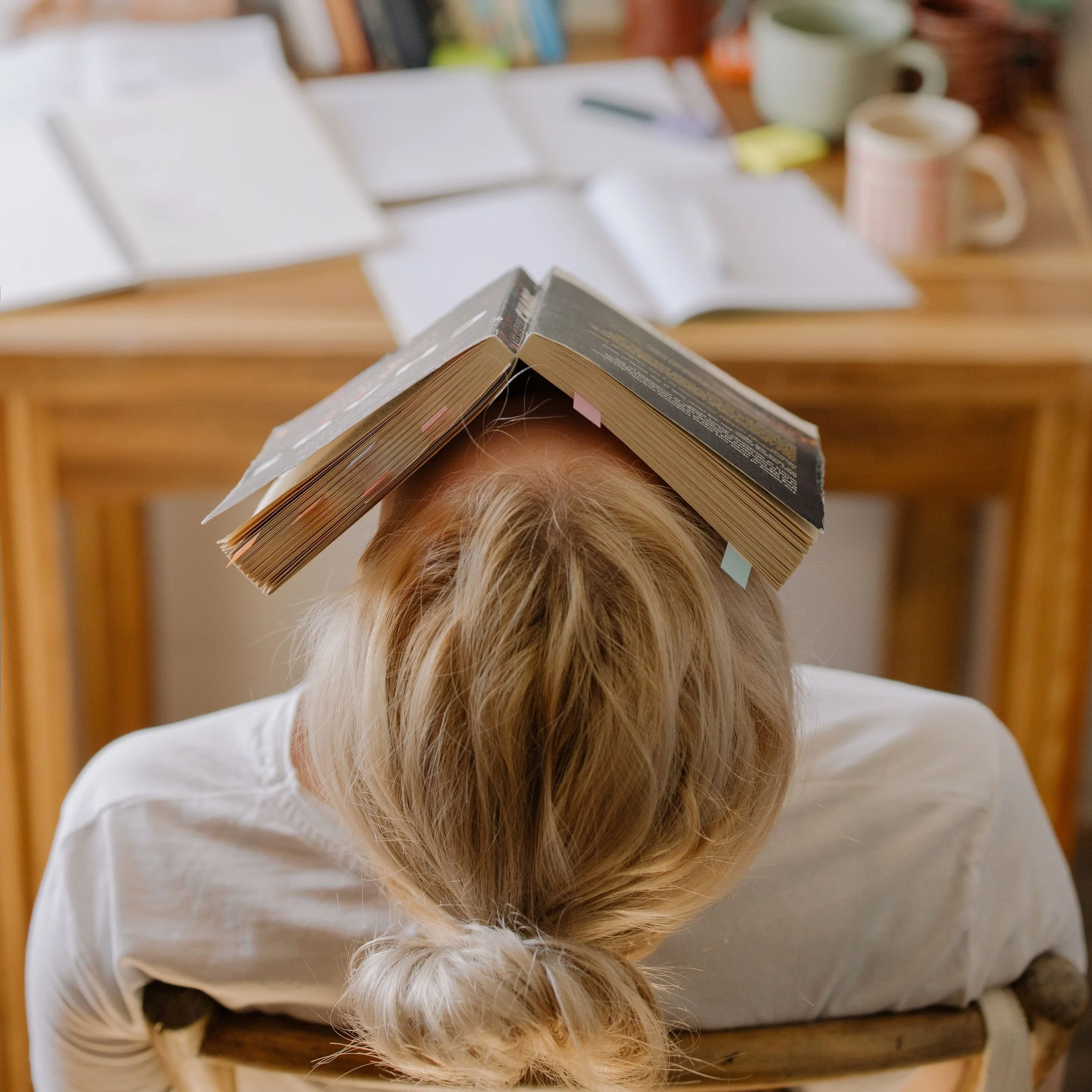 A person with long blonde hair is balancing an open book on their forehead and holding a dog by the neck, against a wooden table with books and mugs in the background.