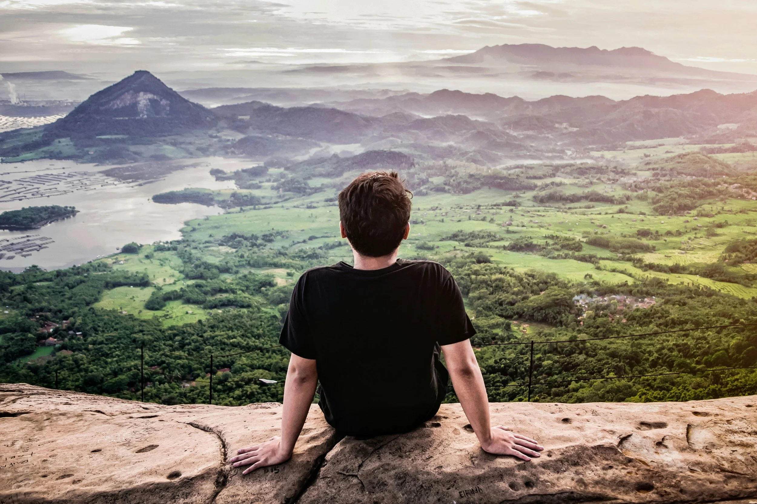 A person sitting on a rocky ledge overlooking a lush, green valley with mountains in the distance, during a cloudy day.