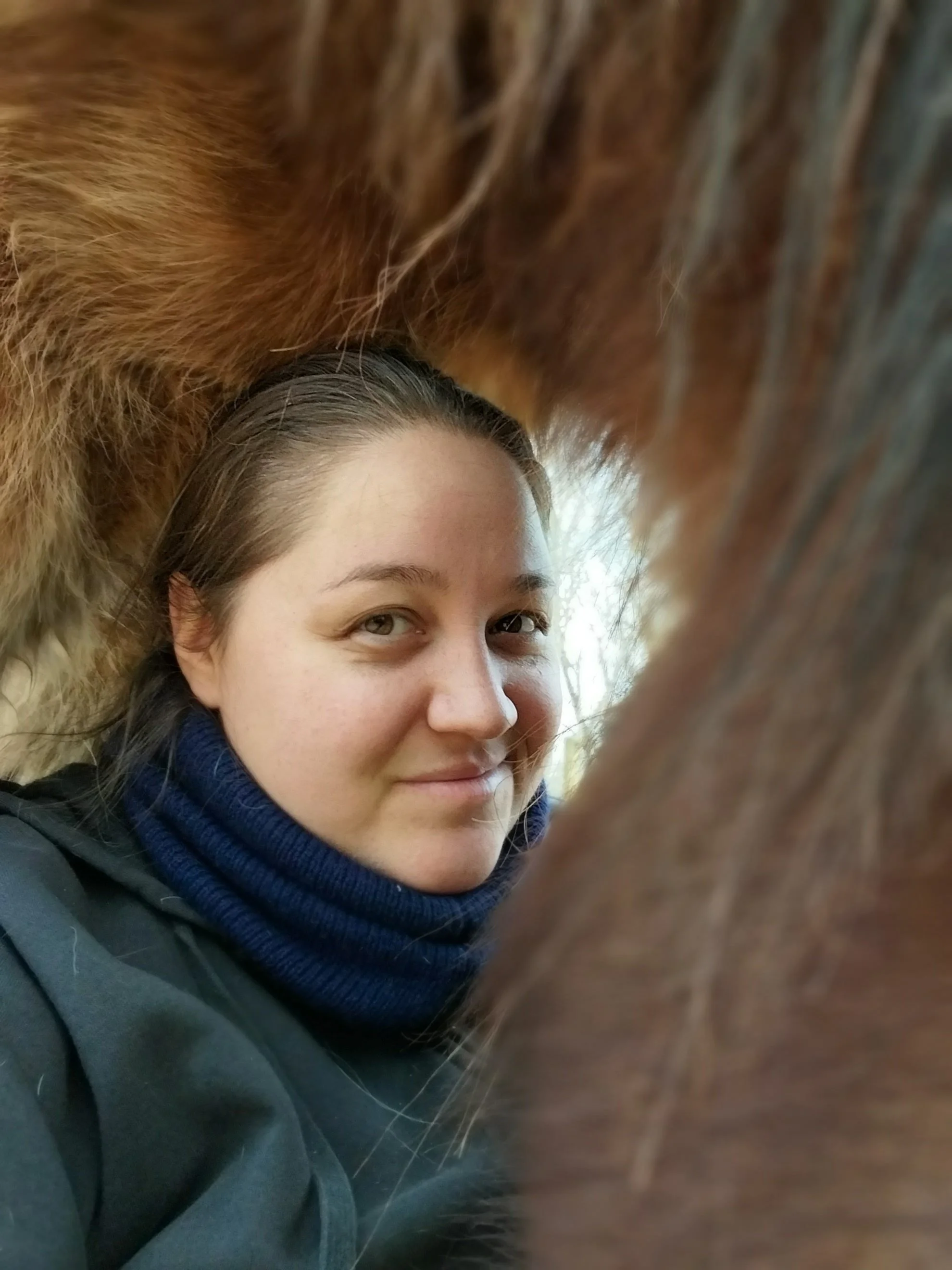 A person with light skin, dark hair, and brown eyes is smiling softly while leaning close to an animal, possibly a horse, with brown fur and a visible mane. The person's face is partially obscured by the animal's fur.