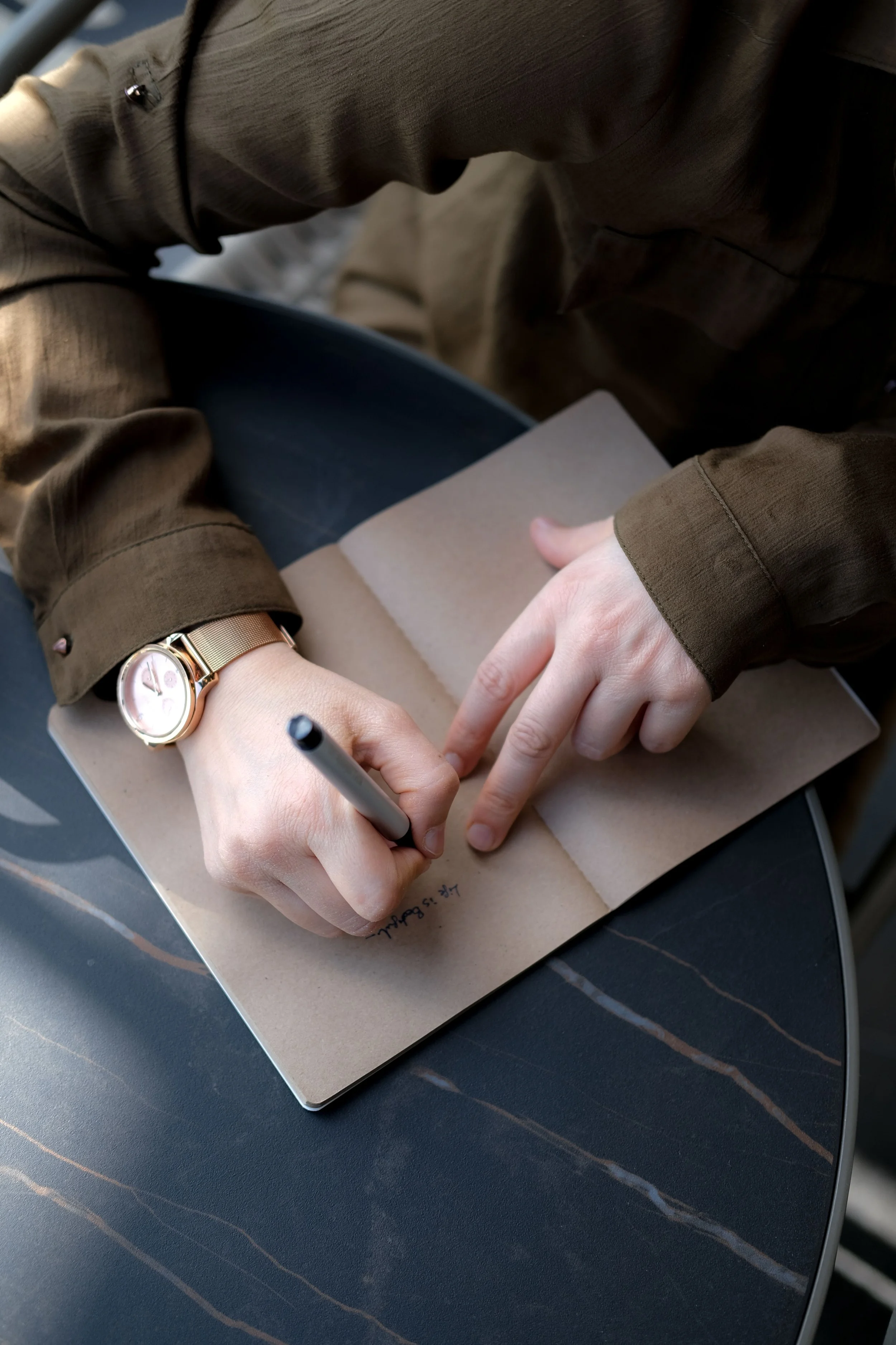 A person wearing a watch and a brown jacket writes in a notebook with a black pen, sitting at a dark table with a subtle striped pattern.