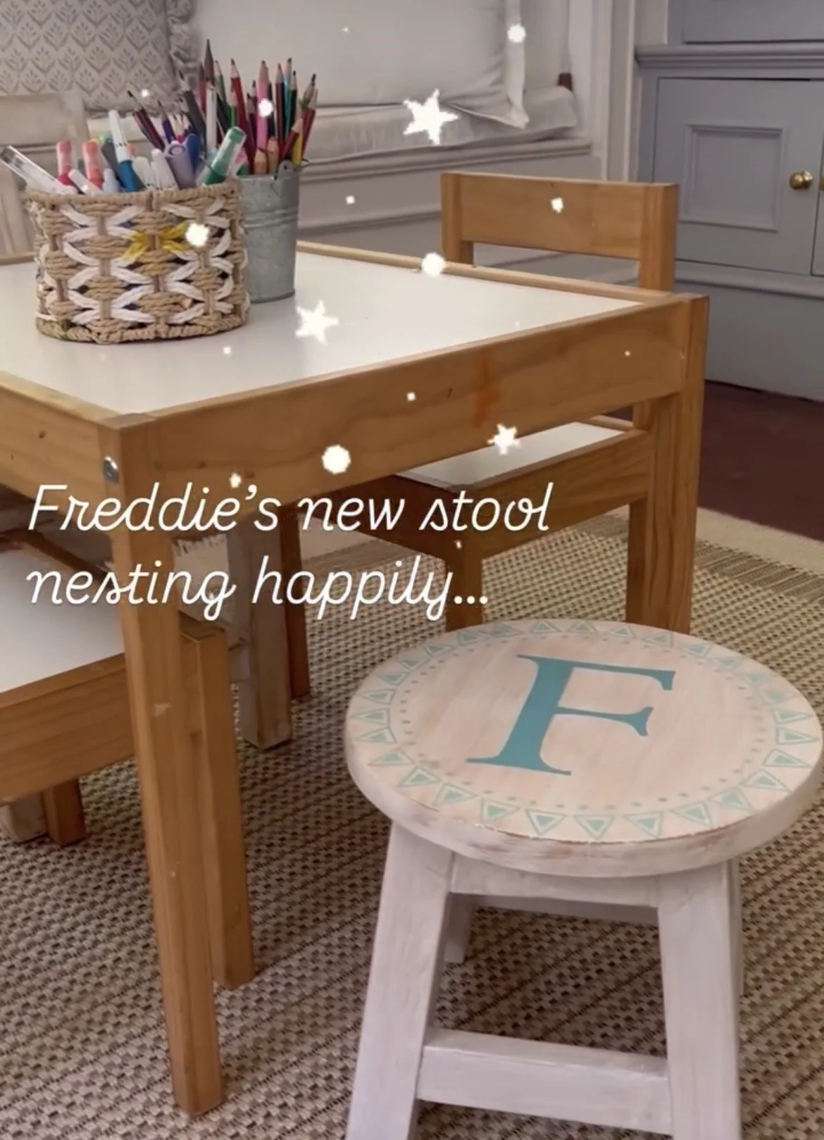 A children's room with a white table holding two containers filled with colored pencils and markers, a small chair with a round top painted with a large blue letter E, and a small bench beneath the table. The room has a gray and white color scheme with a window seat and a gray cabinet. The photo has floating star shapes and the text 'Freddie's new stool nesting happily...' overlaid.