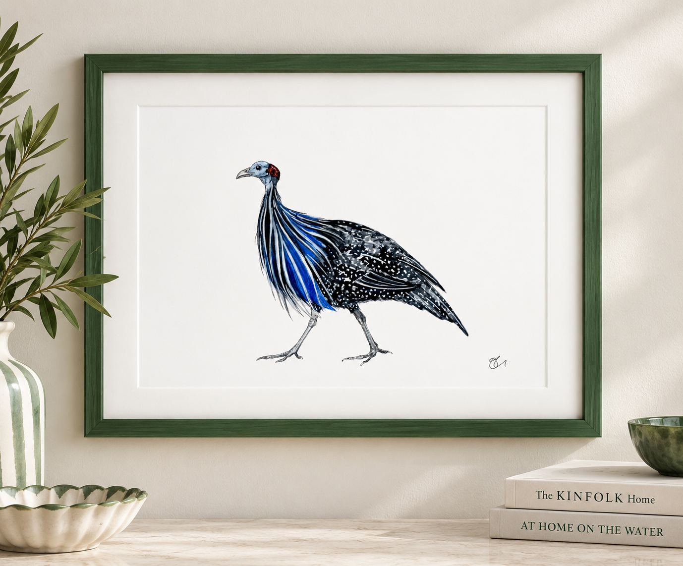 Framed watercolor painting of a bird with black and white spotted feathers and blue streaks on its neck, standing on a white surface, with green plant and books nearby.