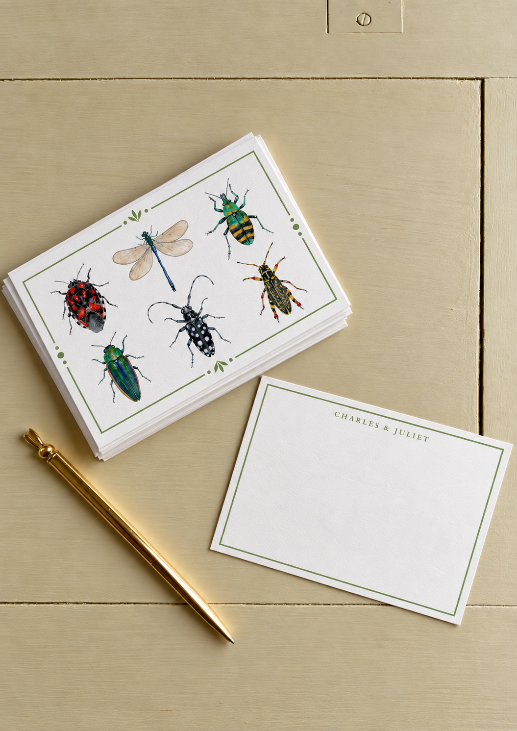 A stack of stationery featuring colorful insect illustrations, with a gold pen and a blank notecard that reads 'Charles & Juliet' on a beige wooden surface.