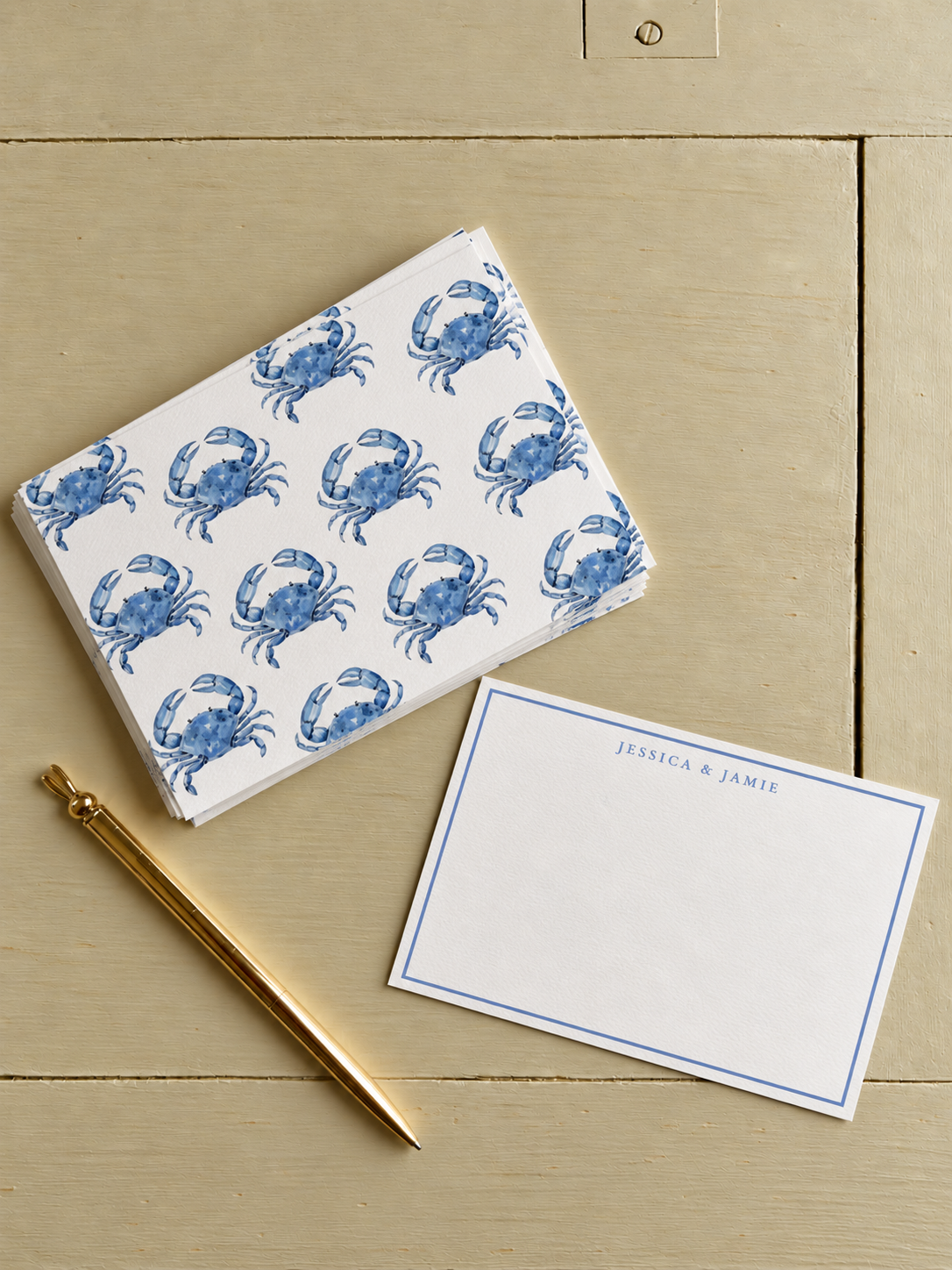 Stack of napkins with a blue crab pattern, a white flat card with blue text 'Jessica & Jamie', and a gold pen on a light-colored wooden surface.
