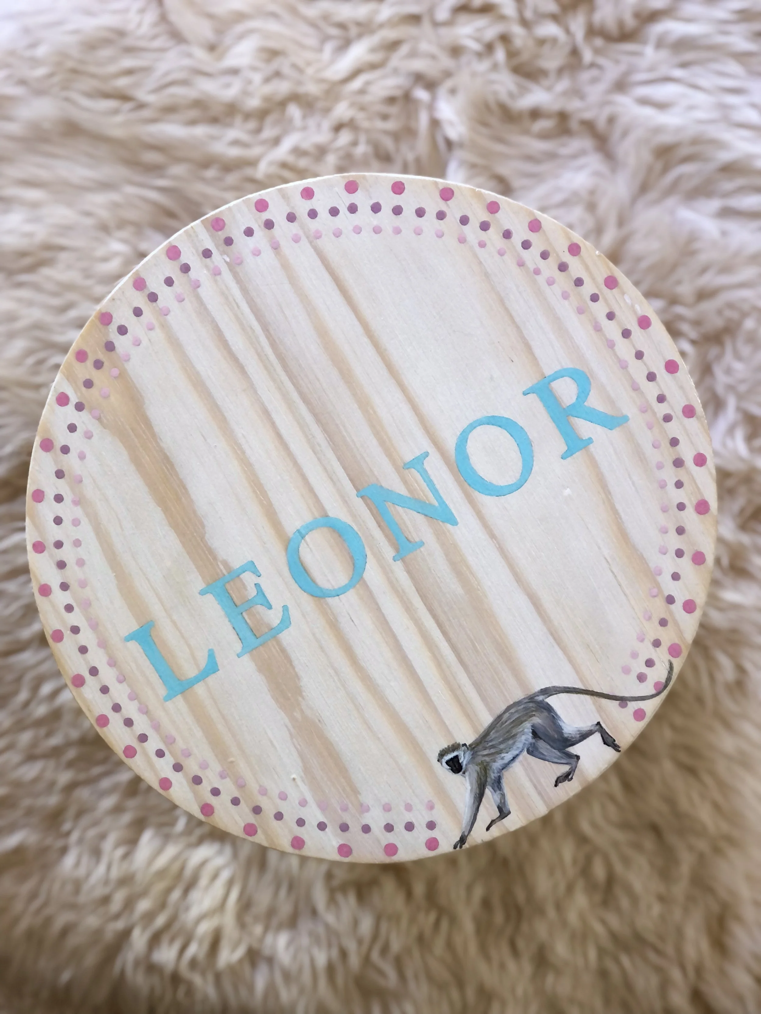 A wooden paddle with the words "LEMON ORE" painted in blue and pink polka dots along the edge, featuring a small illustration of a lemur at the bottom, placed on a beige fuzzy surface.