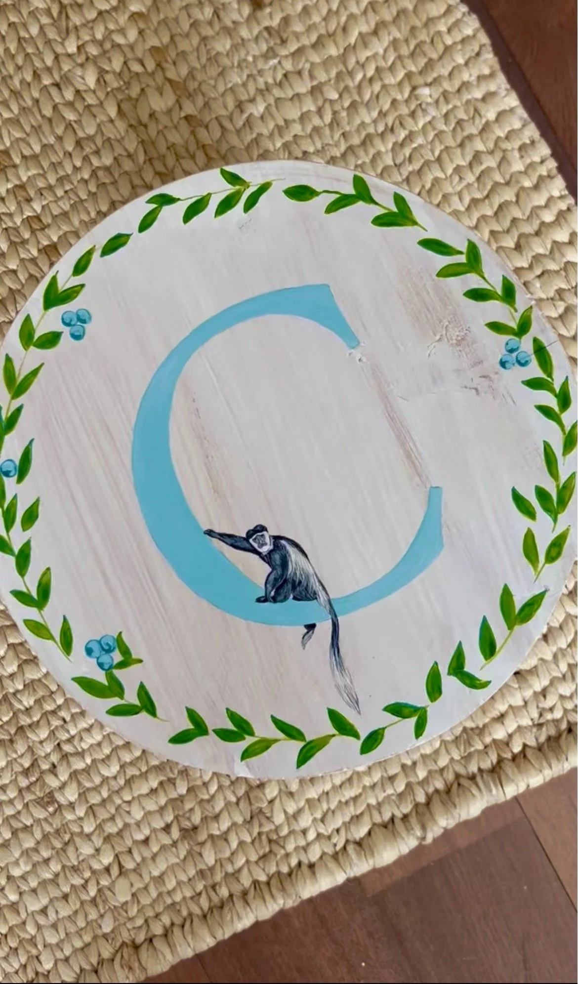 Decorative round placemat with a painted design of a chimpanzee and a large blue letter C, surrounded by a green leafy border and small blue berries, on top of a woven mat.