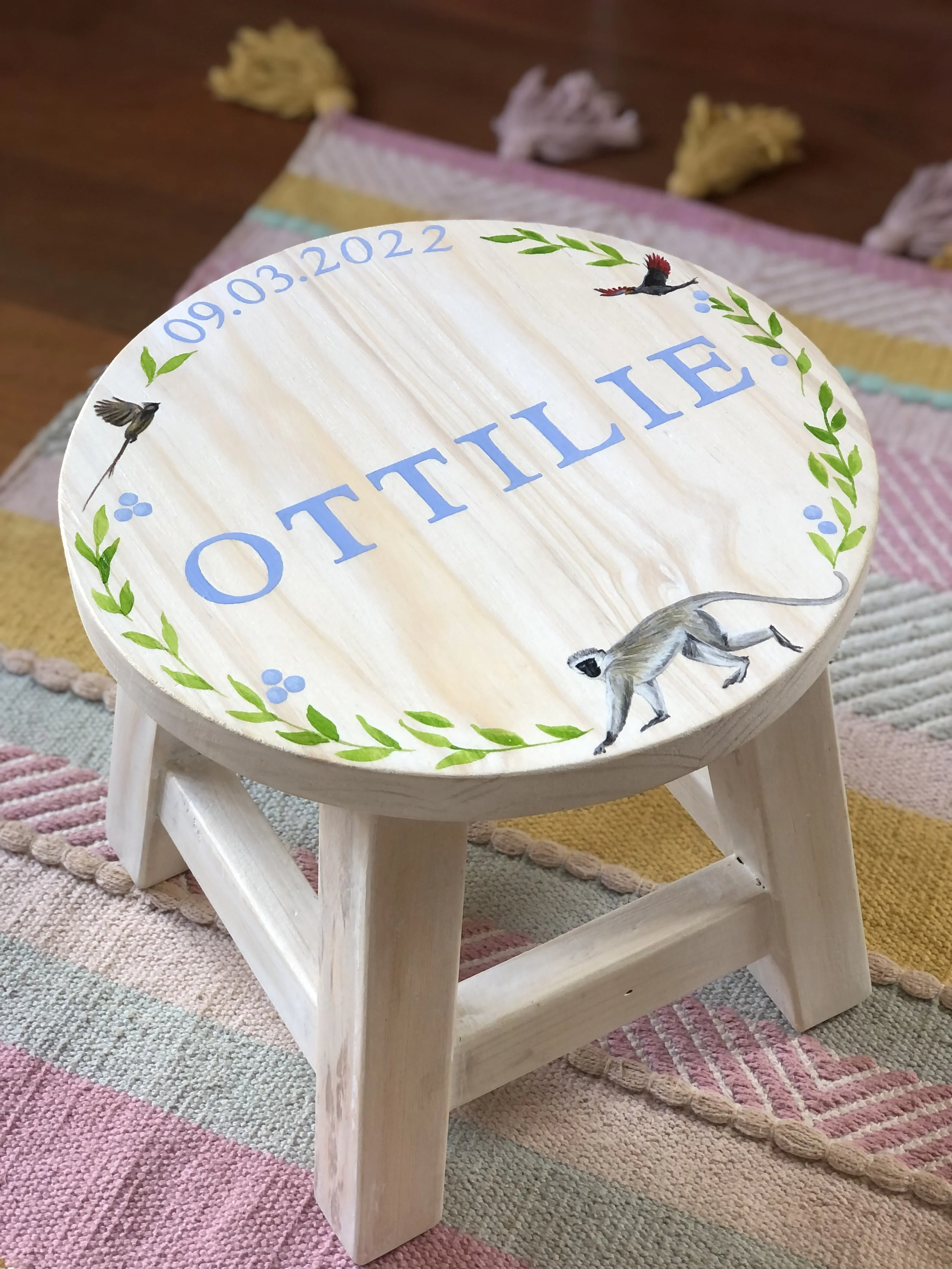 Custom wooden stool with hand-painted design featuring the word "OTITILE," surrounded by birds, leaves, and a monkey, on a colorful woven rug.