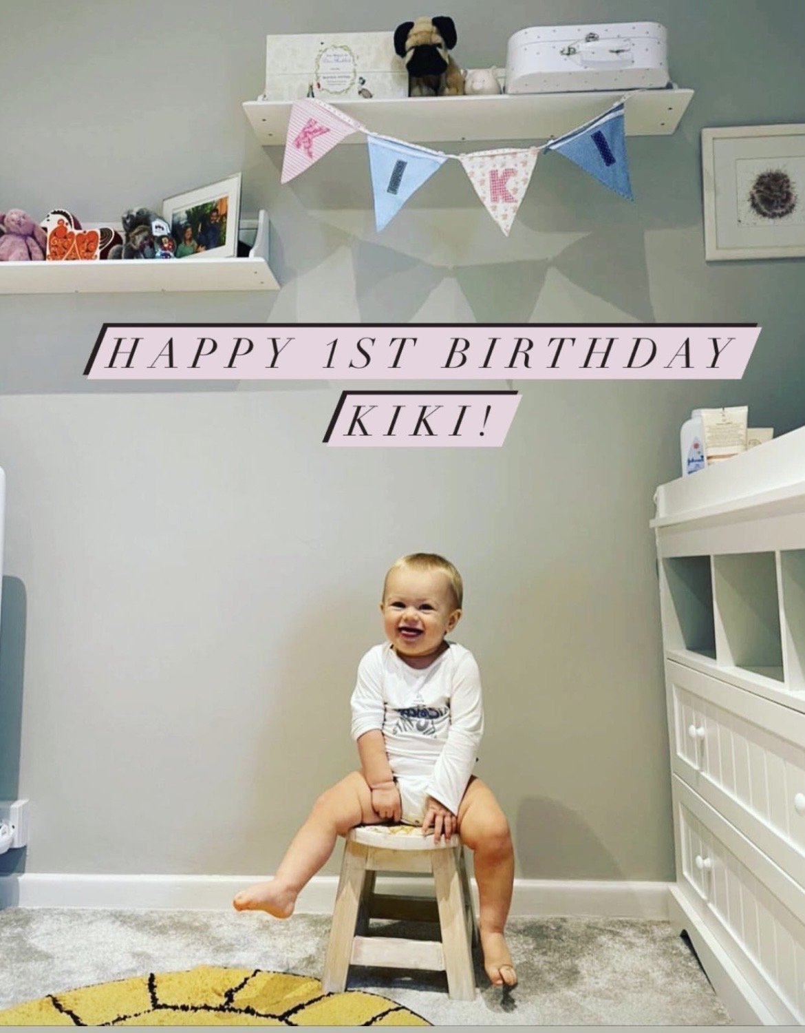 A smiling baby sitting on a small wooden stool with legs spread, in a nursery. The baby is wearing a white long-sleeve shirt. Behind, there is a wall with floating shelves displaying toys and framed photos. Decorative bunting spells out 'KIKI' and 'HAPPY 1ST BIRTHDAY' in pink and blue letters. The room has a gray wall, and a white dresser is visible on the right.
