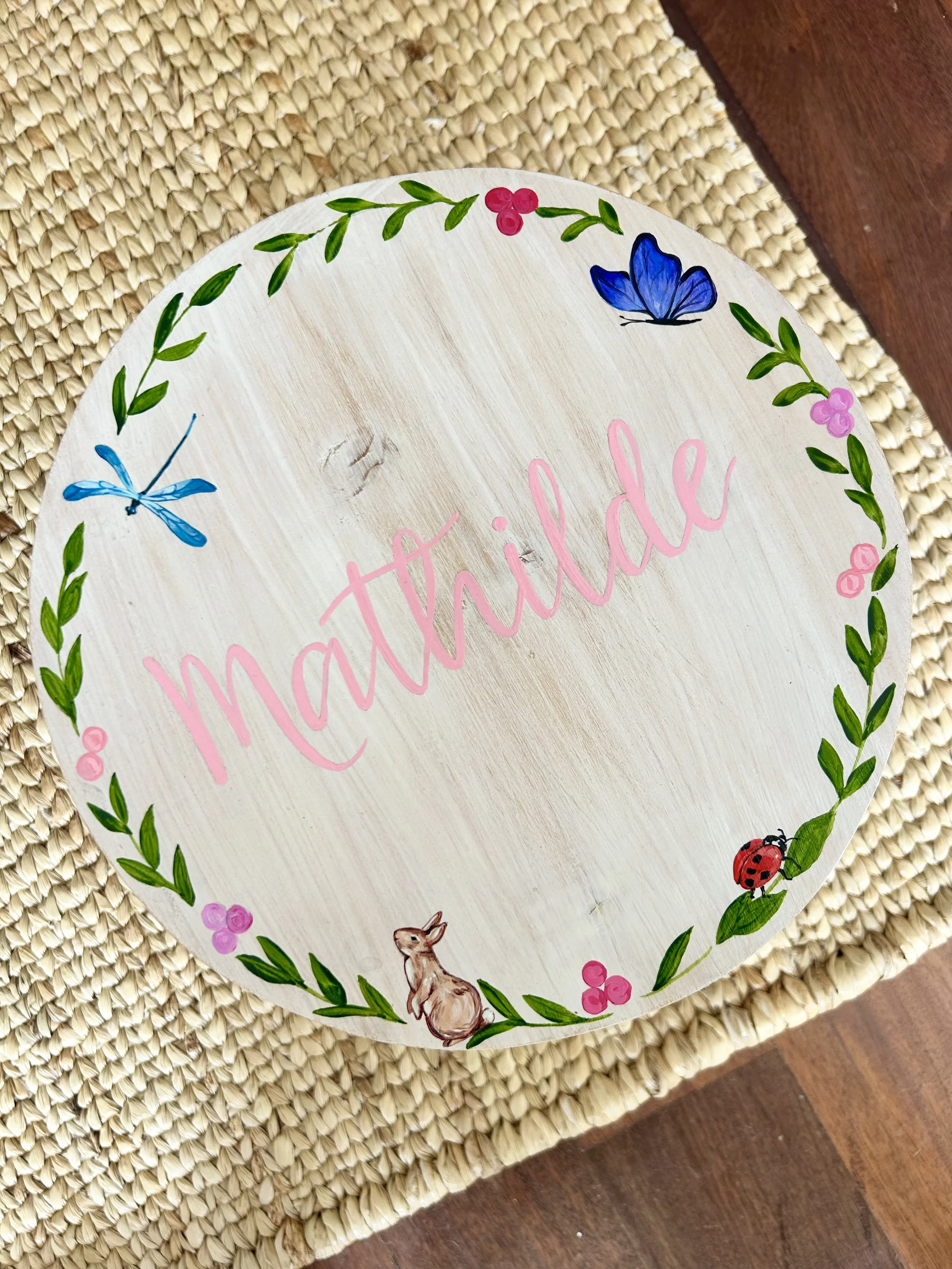 Decorative circular wooden plaque with hand-painted animals and flowers, featuring a blue butterfly, red ladybug, a fawn, pink and purple flowers, and green leafy border, with partial pink unreadable cursive text in the center.