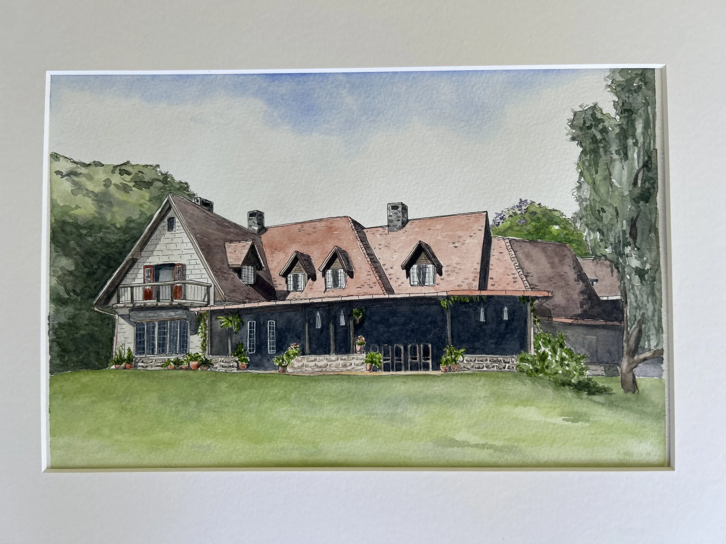 Watercolor painting of a house with a steep roof, dormer windows, and a small balcony, surrounded by green trees and lawn under a blue sky.