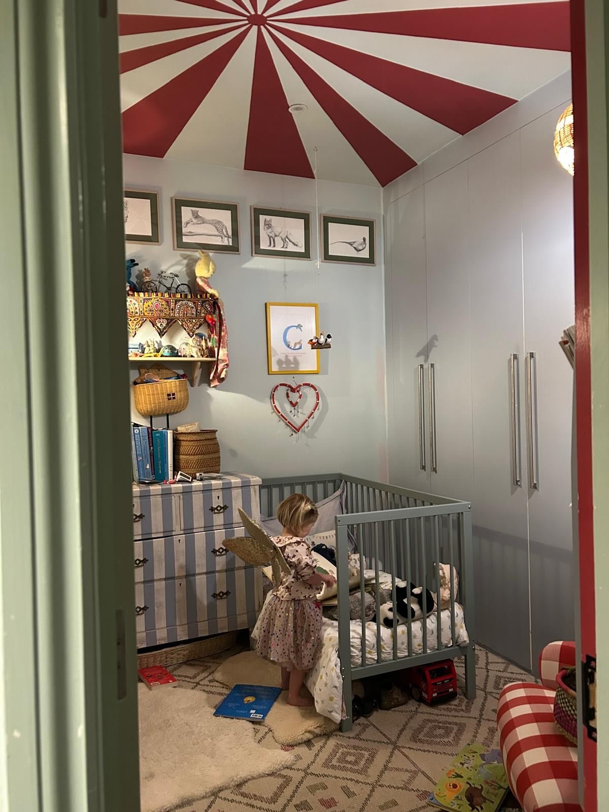 A young girl standing by a gray crib in a colorful nursery, playing with plush toys. The room has a red and white striped tent-like ceiling, framed animal illustrations on the wall, a white wardrobe with long handles, and a striped dresser with books and baskets on top. The decor includes a heart-shaped wall hanging and a framed letter 'C' with small decorative shoes underneath.