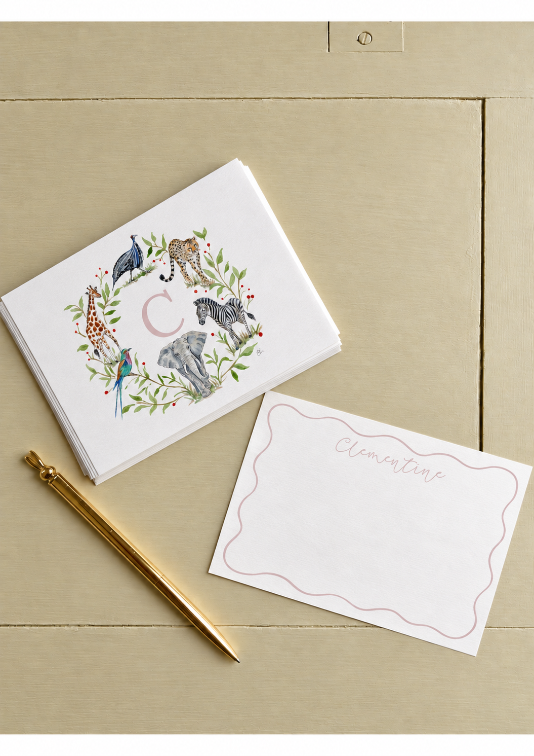 A set of illustrated alphabet cards with jungle animals arranged in a circle around the letter 'C', placed on a beige surface along with a white card with the handwritten name 'Clementine' and a gold pen.