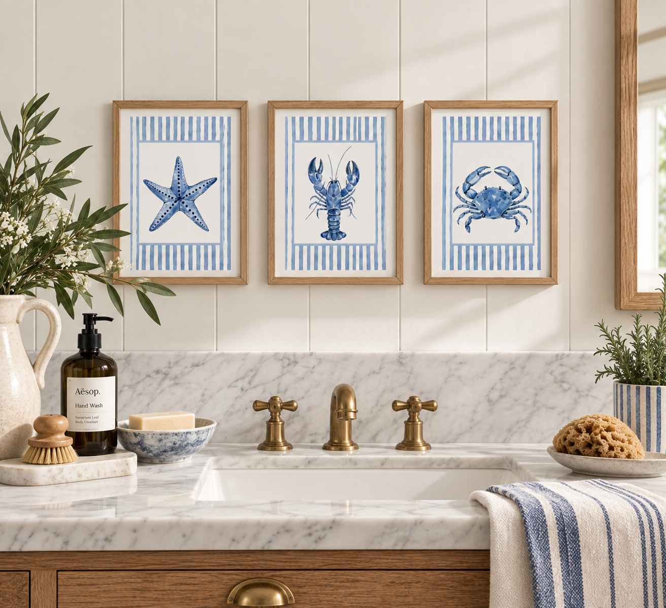 Bathroom sink with marble countertop, brass faucet, and framed beach-themed artwork featuring a starfish, lobster, and crab on the wall. Decor includes a plant, soap dispenser, brush, soap, sponge, and a striped towel.