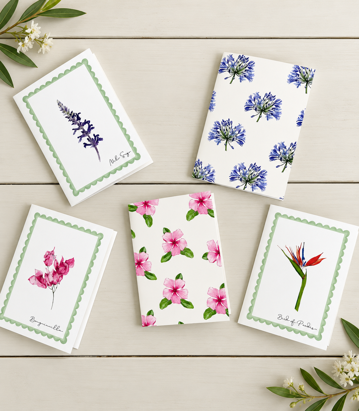 Set of five greeting cards with floral designs, including lavender, blue agapanthus, pink mandevilla, pink impatiens, and red bird of paradise, arranged on a white wooden surface with small sprigs of flowers and leaves.