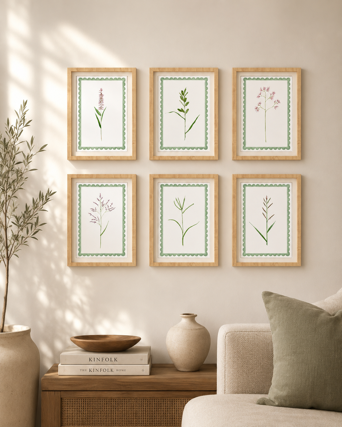A living room wall decorated with six framed botanical prints arranged in two rows of three. Each frame contains a simple plant illustration with a green border and light wood frame. To the left, there is a potted plant. Below the prints, on a wooden side table, are two books titled 'Kinfolk' and a decorative bowl. A beige sofa with a green cushion is visible in the bottom right corner.