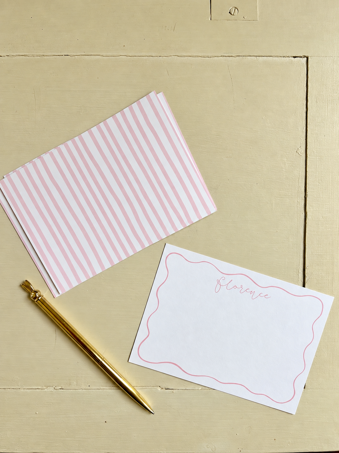 A gold pen, a pink and white striped card, and a white card with pink border and the name Florence written on it, all on a beige wooden surface.