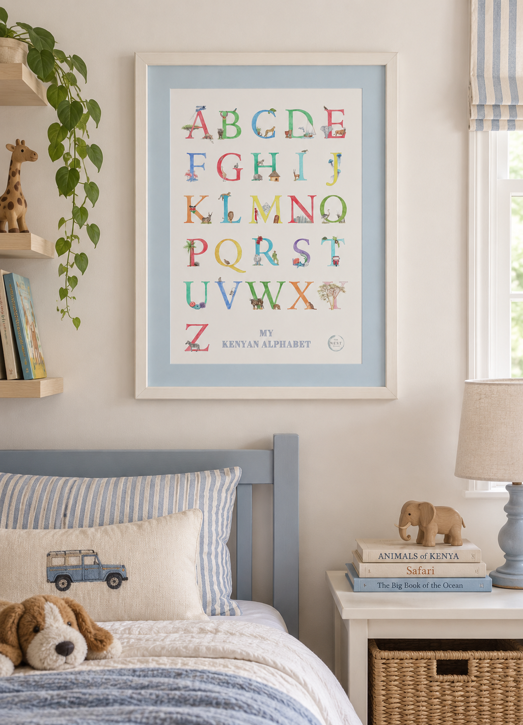 Kids' bedroom with a framed poster of the Kenyan alphabet, a bed with paw print and car pillows, a nightstand with books about animals and a wooden elephant, a lamp, and a window with blue and white striped curtain.
