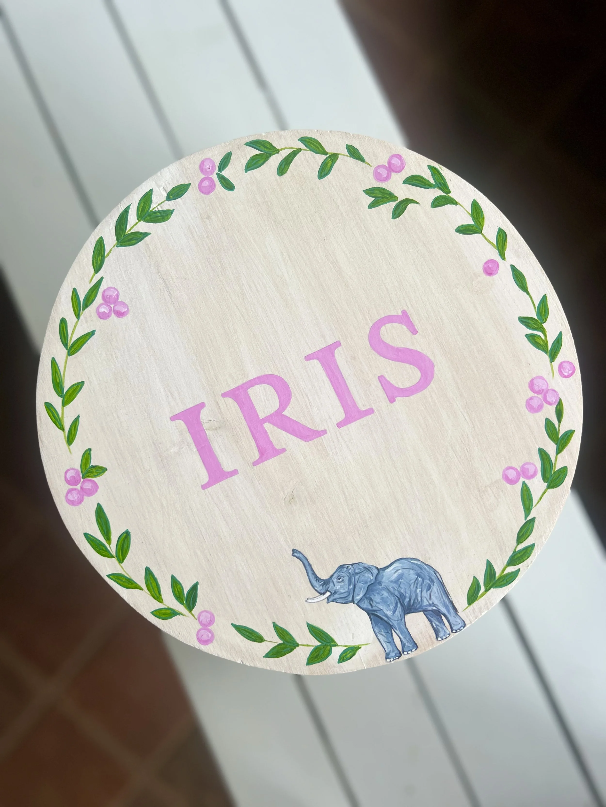 A circular sign decorated with a green leafy border and pink berries, featuring the word 'TRIS' in pink letters and an illustration of a blue elephant at the bottom.