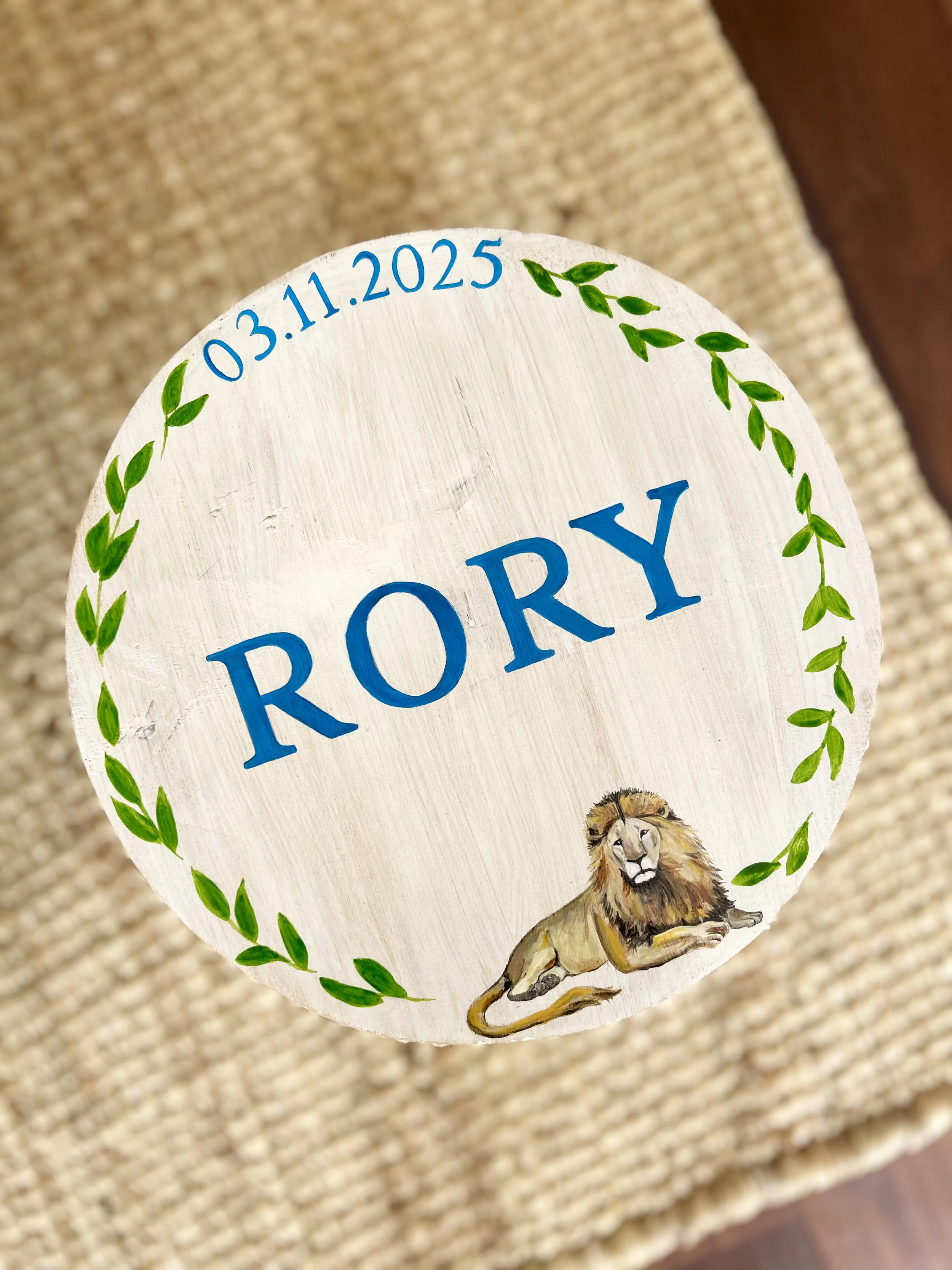 A painted wooden sign with a lion illustration and green leaf border, displaying the date 03.11.2025 and the word 'ROY'.