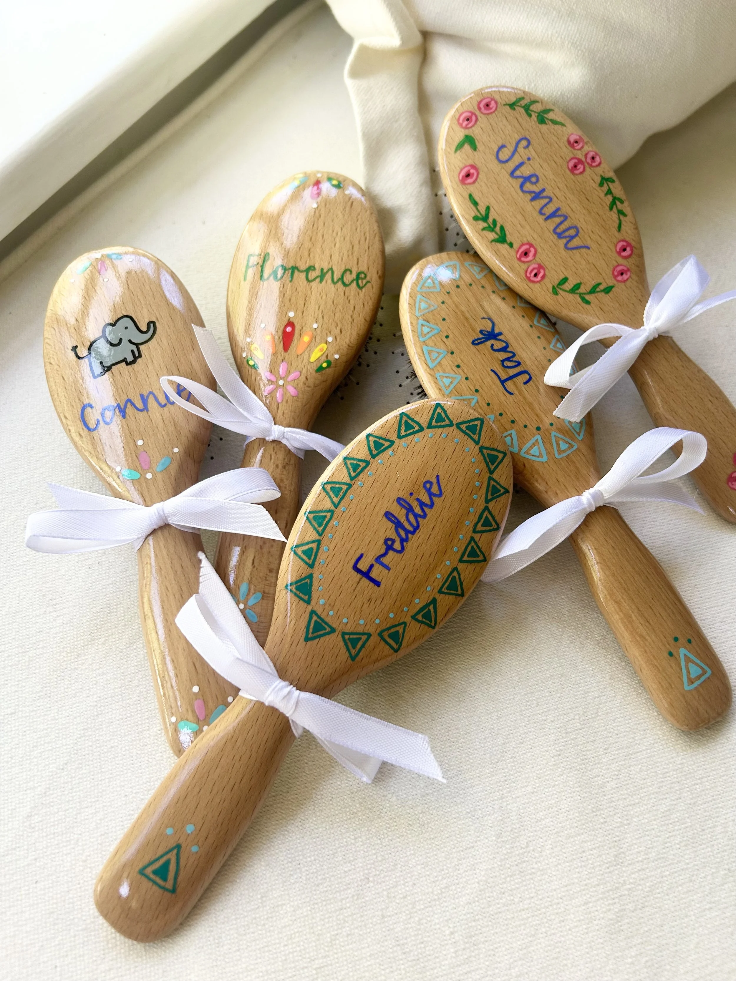 Hand-painted wooden spoons with names and decorative patterns, tied with white ribbons, resting on a beige fabric surface.