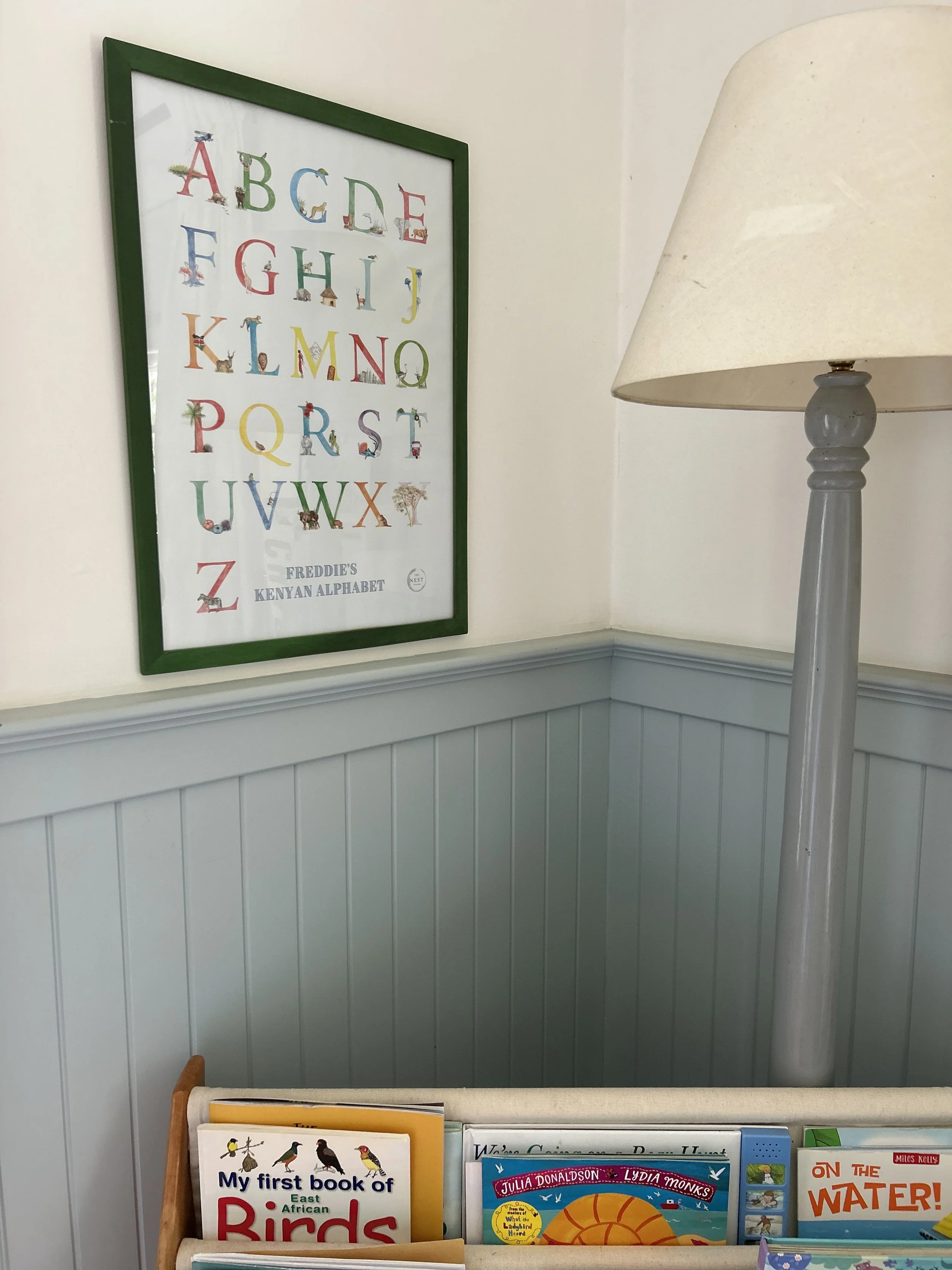 A framed alphabet poster on a wall with colorful letters and animal illustrations, next to a bookshelf with children's books.