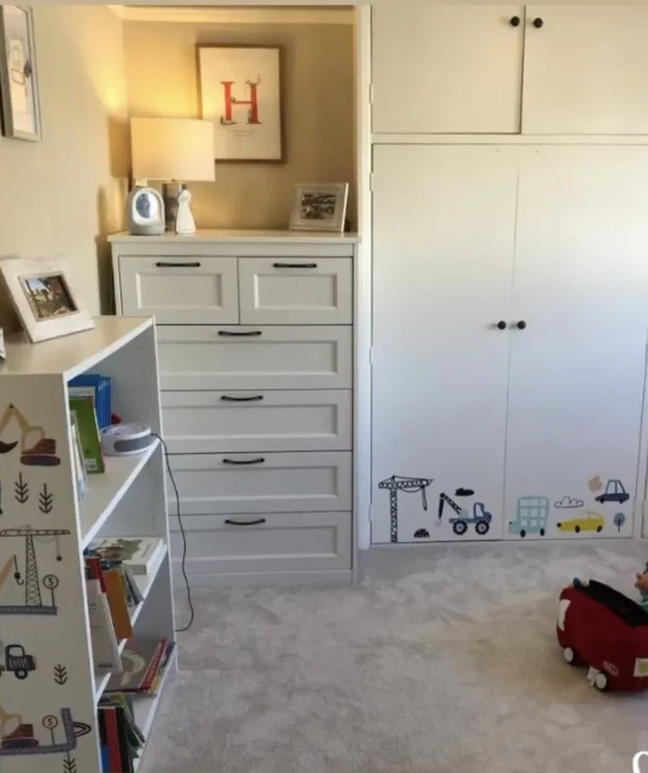 A corner of a child's room with a white dresser, a lamp, framed pictures, a bookshelf filled with books, and a closet with construction-themed stickers at the bottom. There is a toy car on the floor.